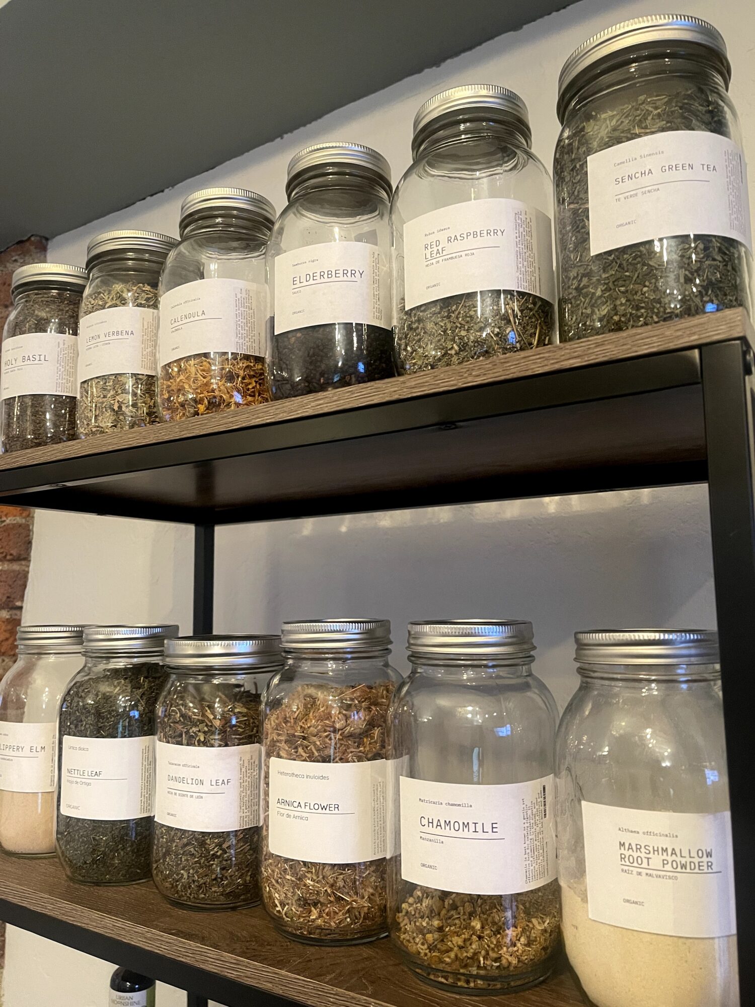 Two shelves with glass jars containing herbs, spices, and powders, some labeled, in a room corner.