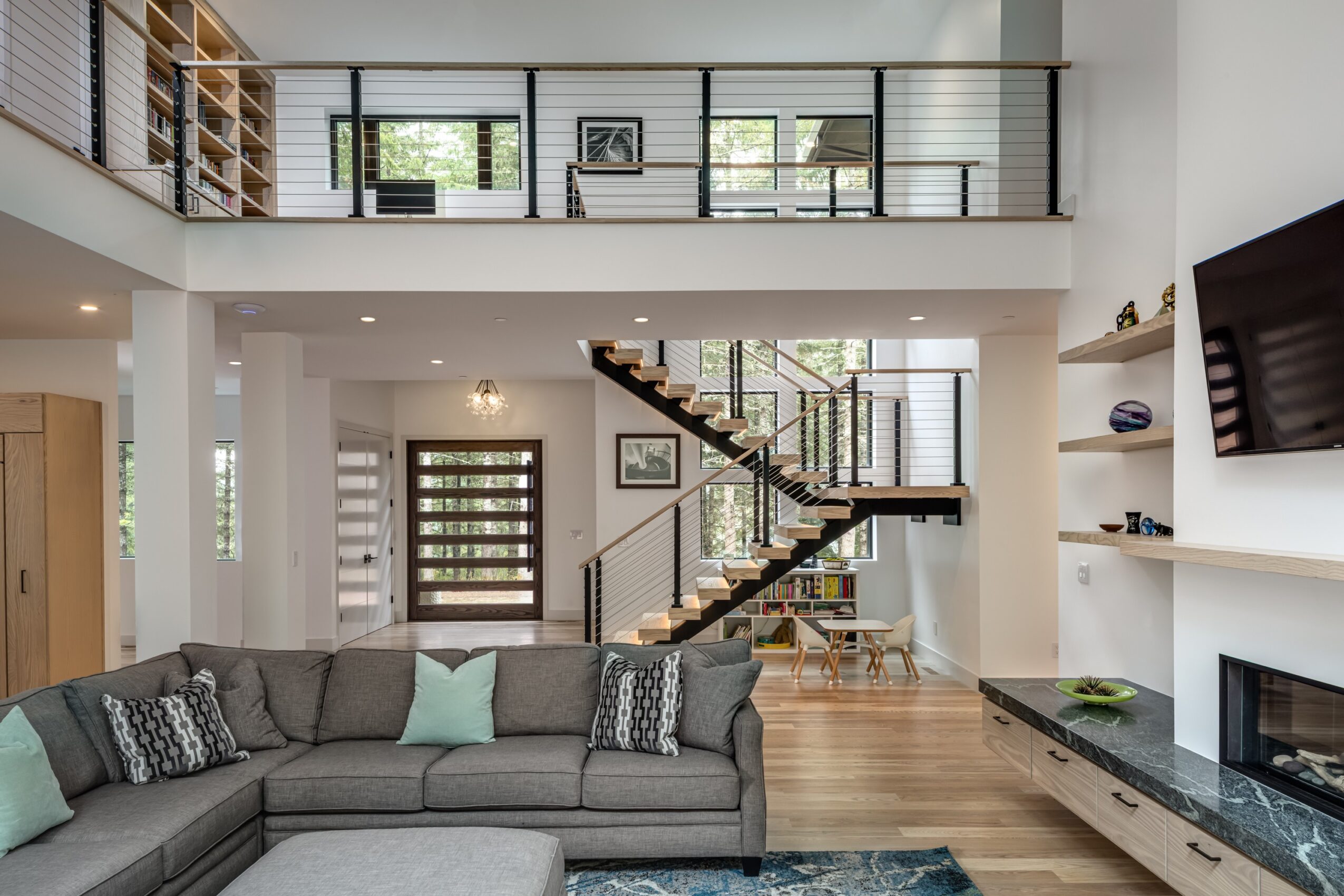 Living room with gray sectional sofa, staircase, and open second floor with railing, TV on wall, and wooden flooring.