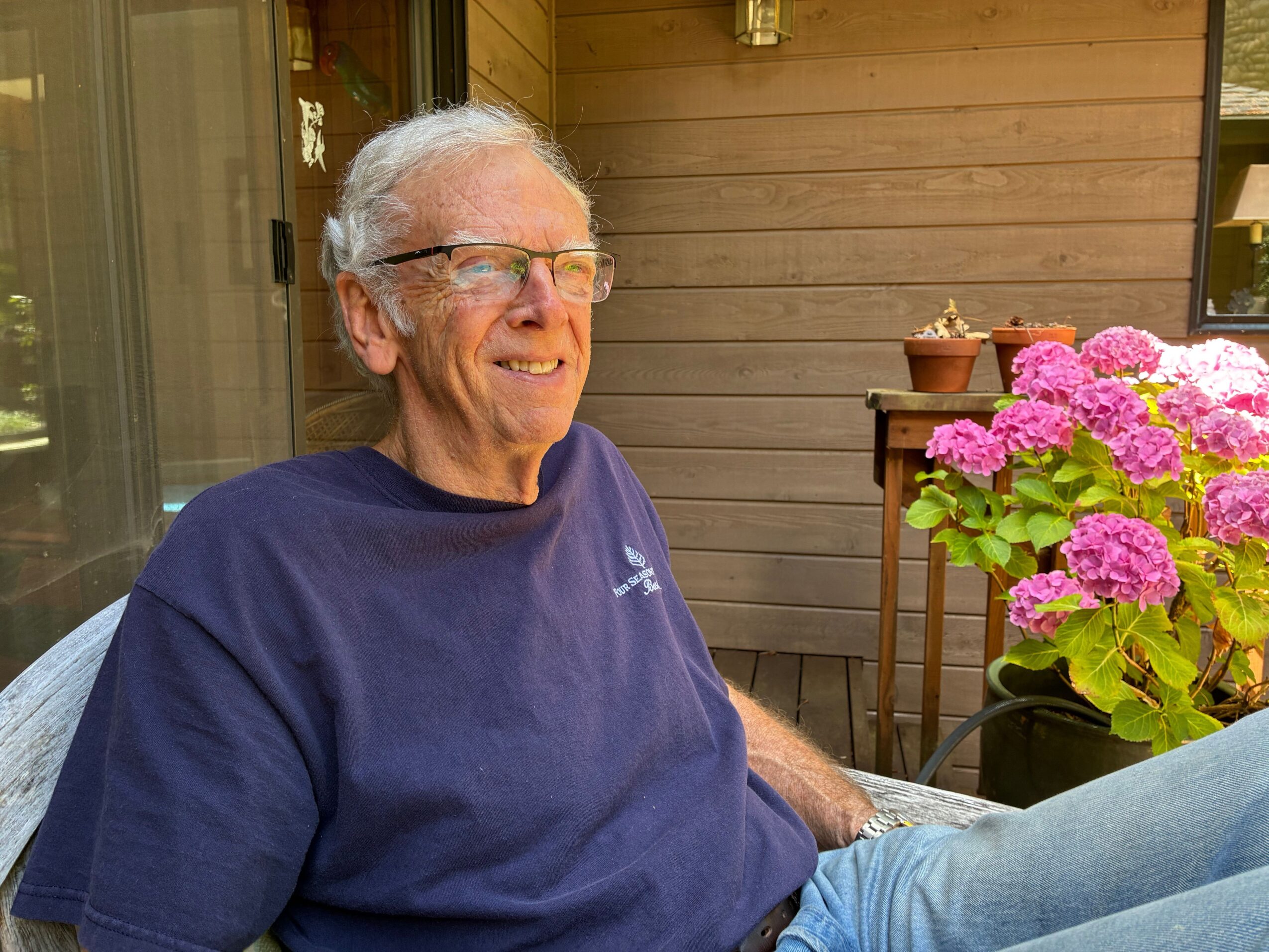 Older man with glasses and gray hair smiling, wearing a navy blue shirt, sitting outdoors near pink flowers and wooden fence.