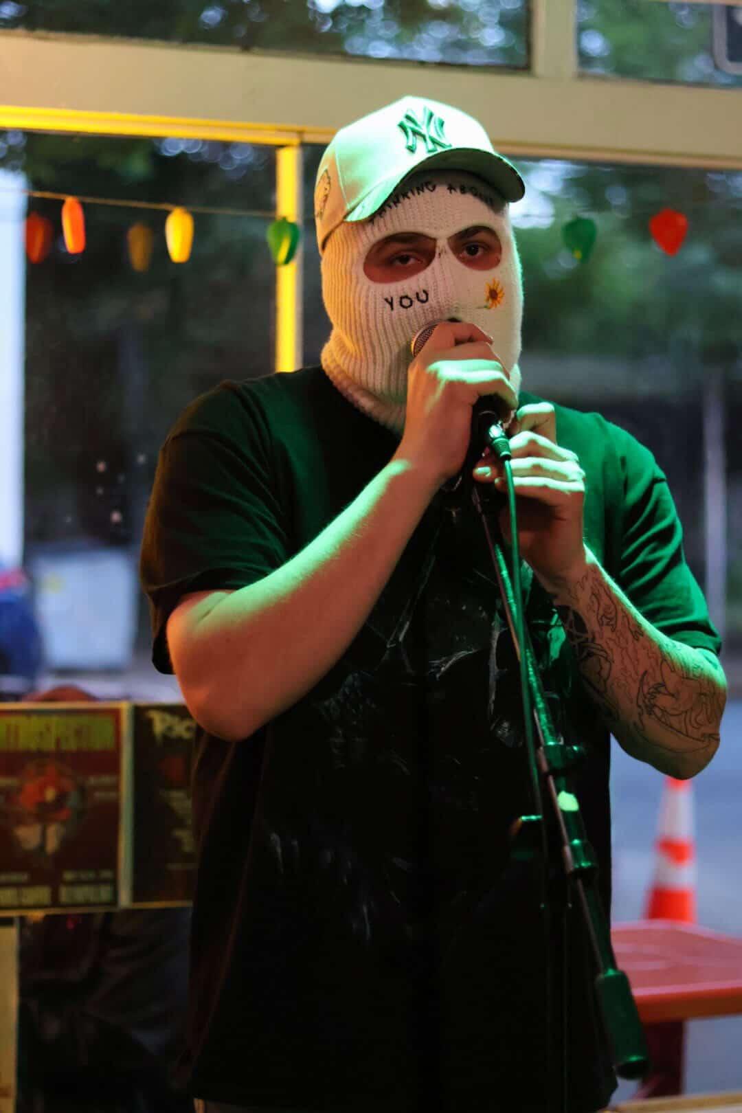 Person wearing a mask with a face design, holding a microphone, in a casual setting with colorful lights in the background.