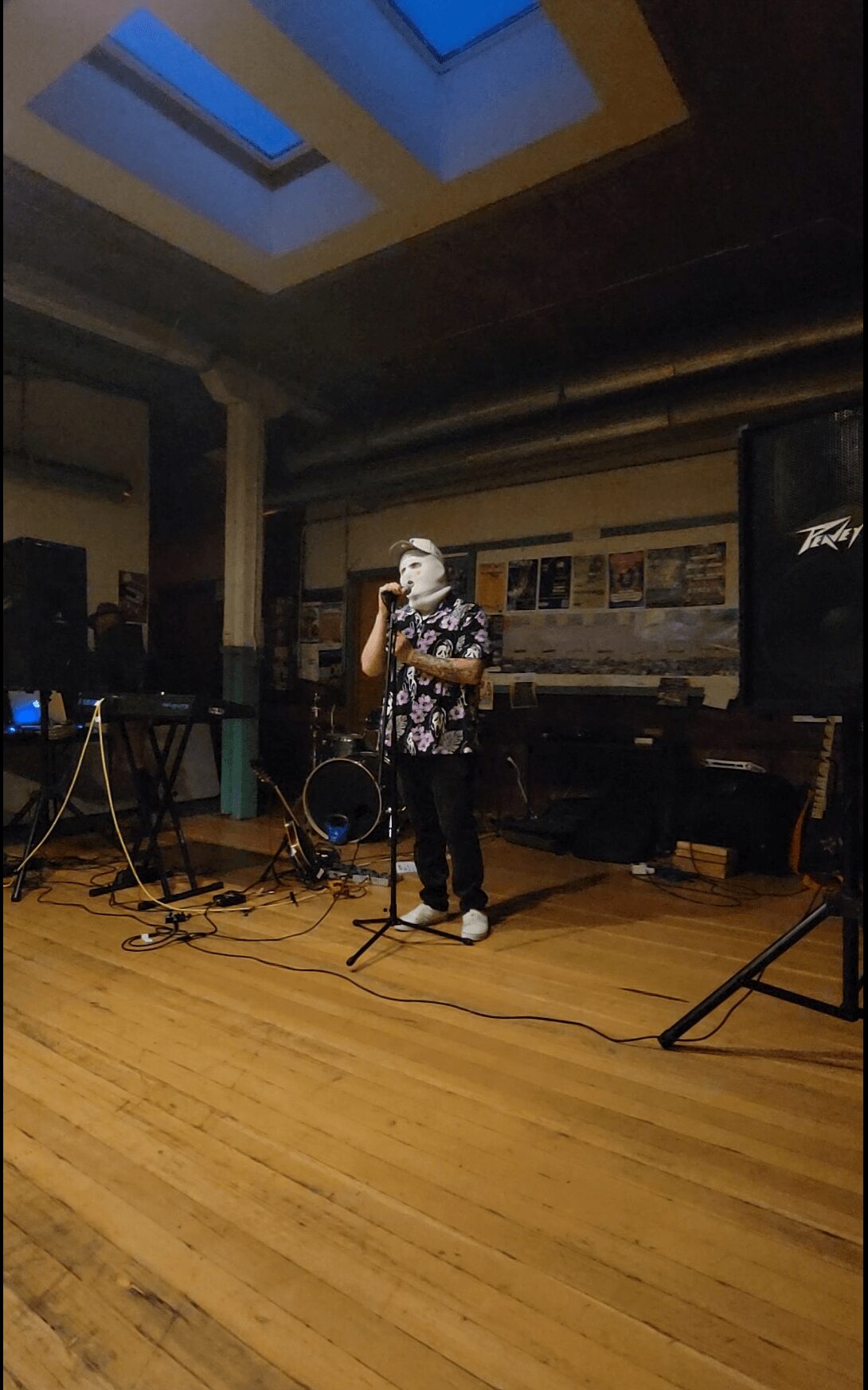 Person wearing a mask and patterned shirt singing into a microphone on stage with musical equipment around.