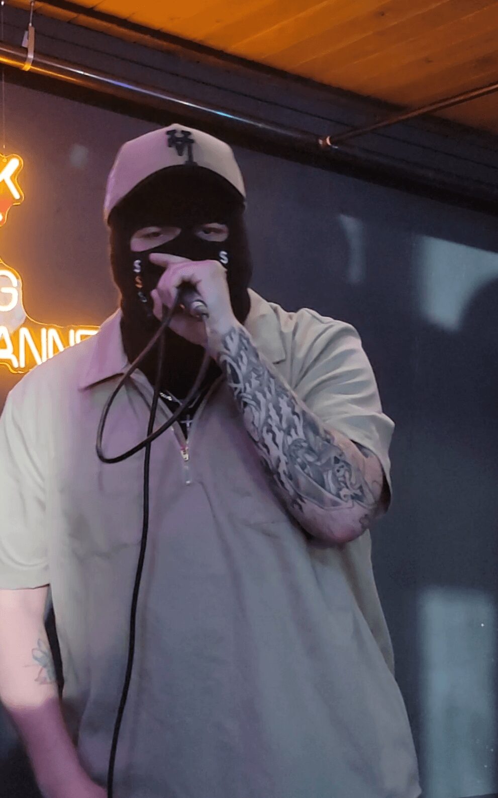 Person wearing a white cap and black mask, holding a microphone, with tattoos on arm, in a dimly lit setting