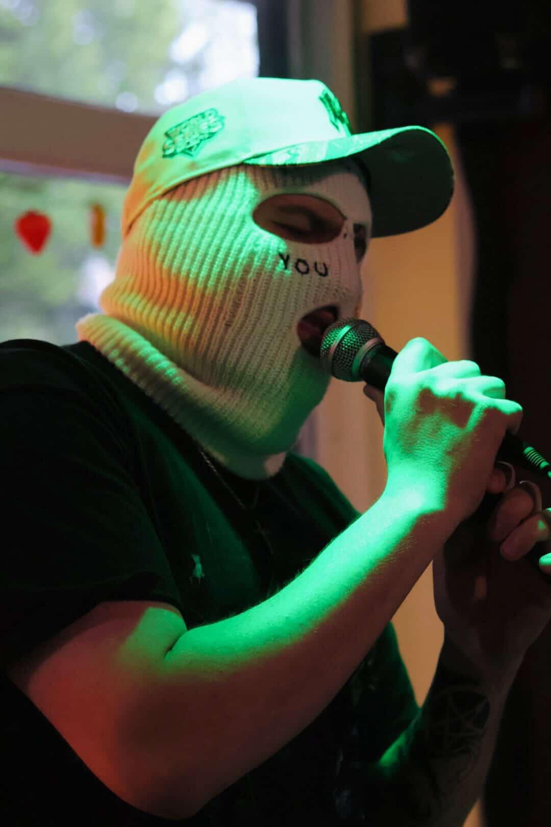 Person wearing a white knit hat and mask, holding a microphone, illuminated by green light, indoors.