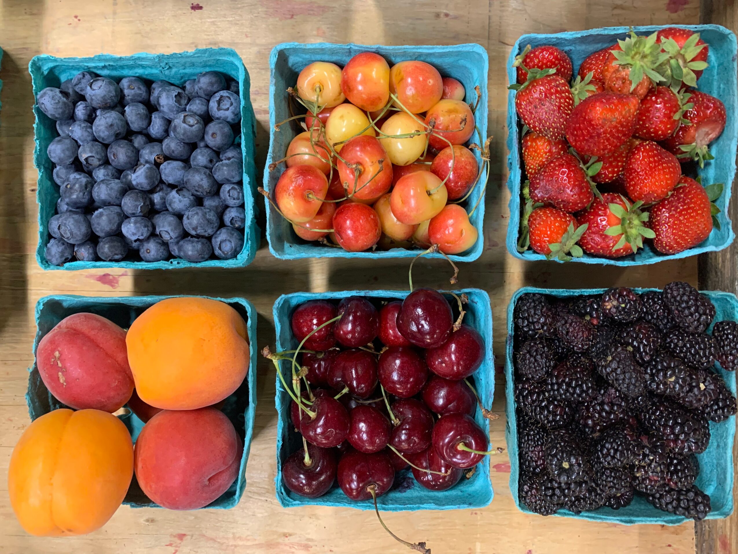 Six baskets of fresh berries and fruits including blueberries, gooseberries, strawberries, peaches, cherries, and blackberries, arranged on a wooden surface.