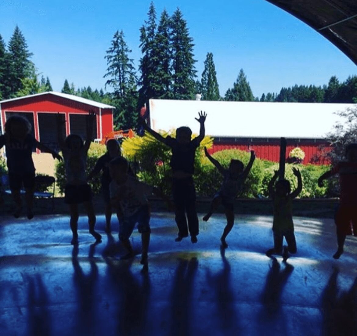 Children playing and jumping indoors with a view of trees and red buildings outside, sunlight streaming in.