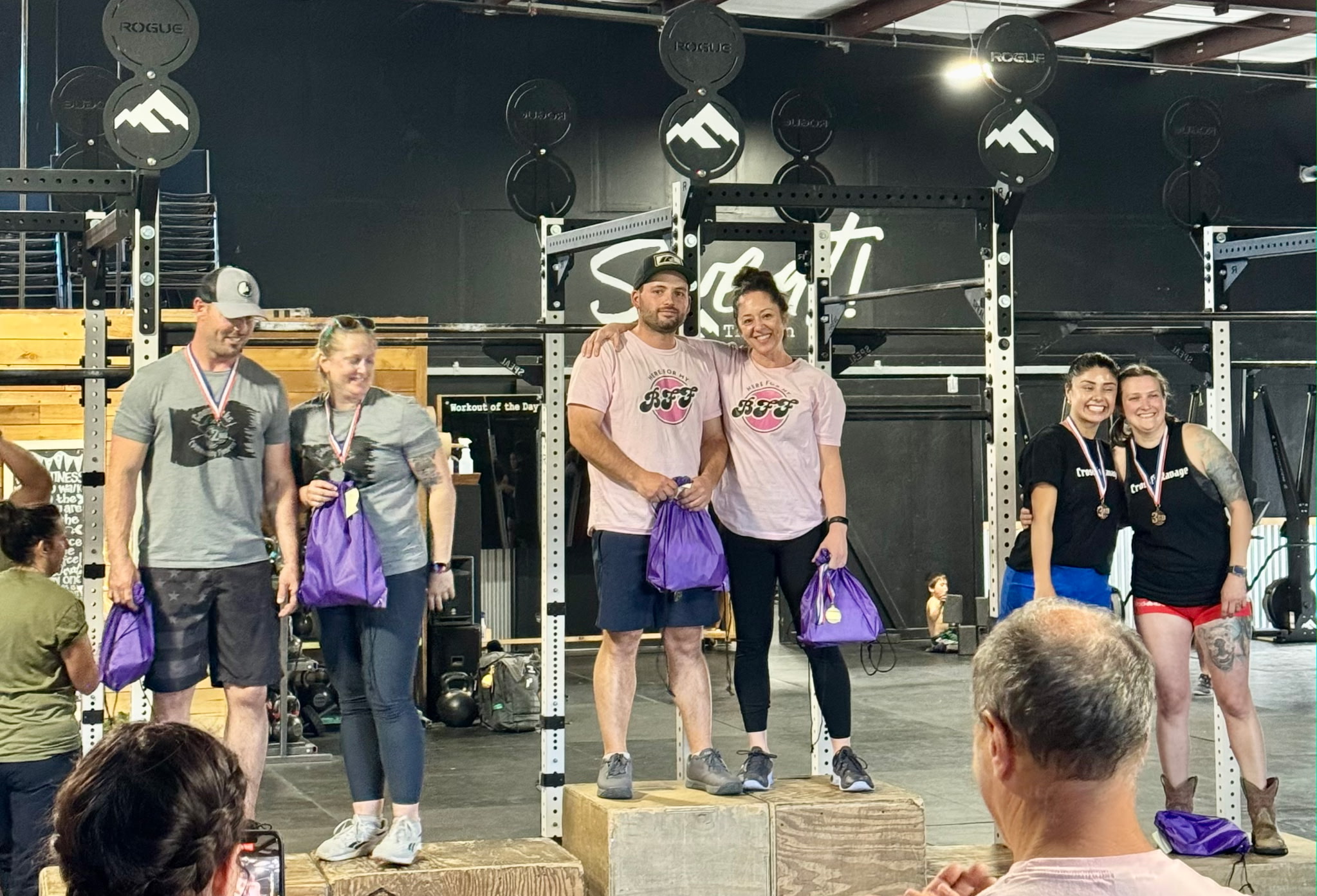 Group of people on stage with medals and purple bags, in a gym setting with equipment in background.