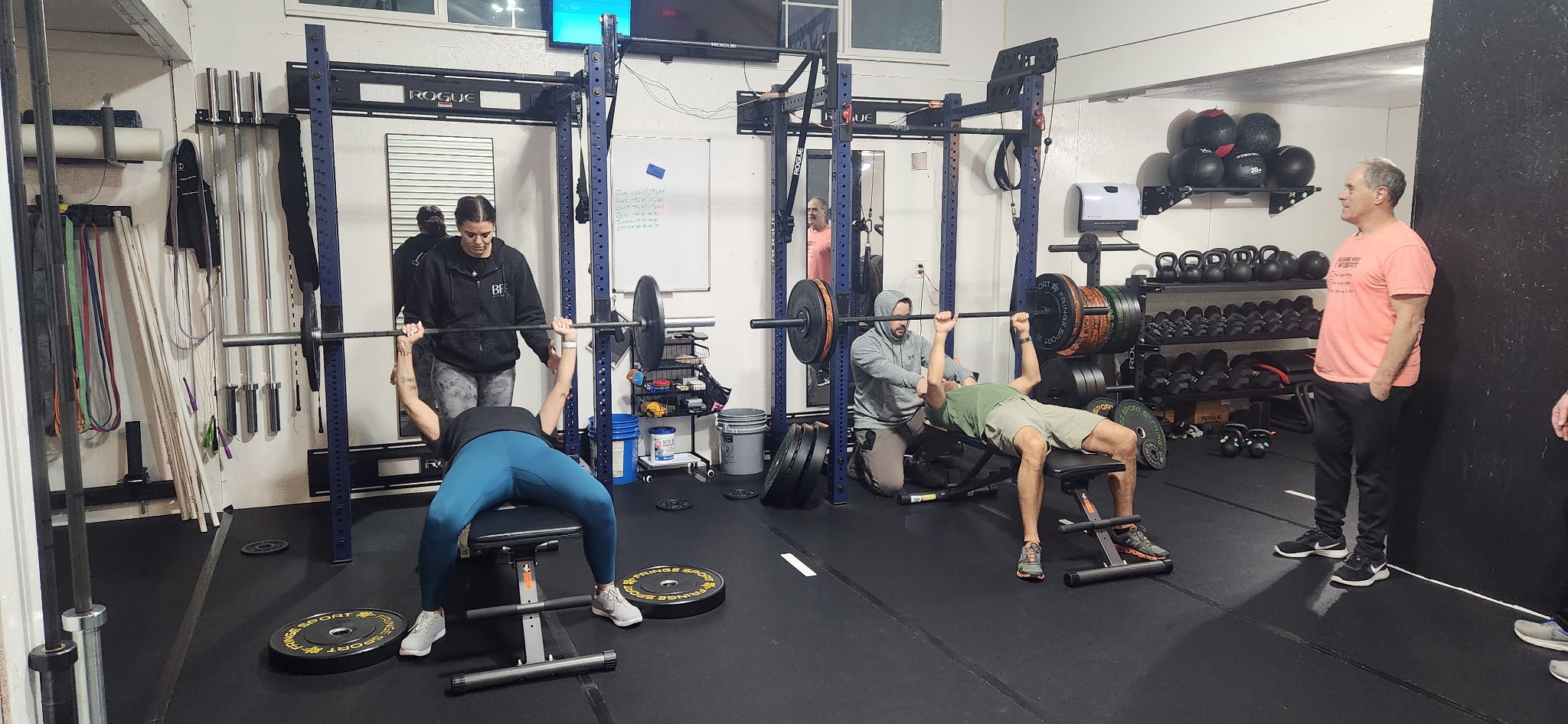 People exercising with weights and benches in a gym, with equipment on walls and floor, and a person standing nearby.