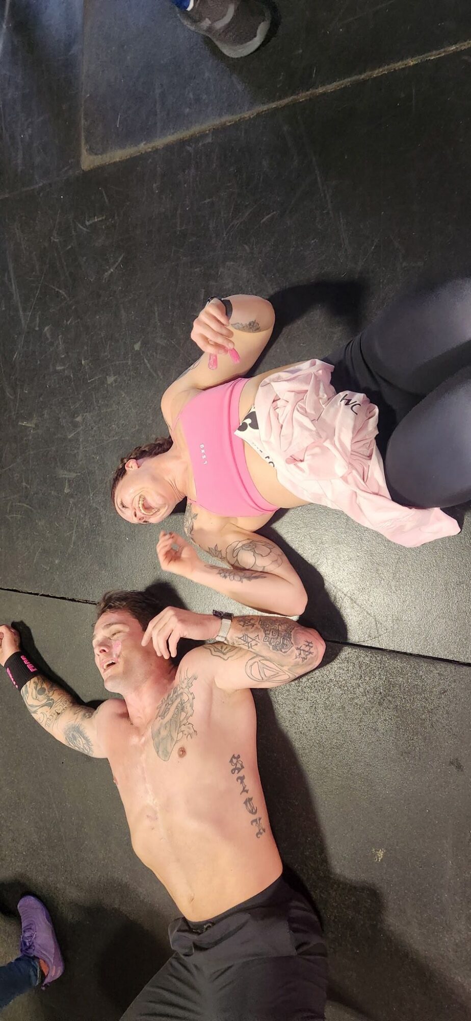 Two people lying on the floor, one with tattoos and shirtless, the other in a pink top and light-colored shorts, resting.