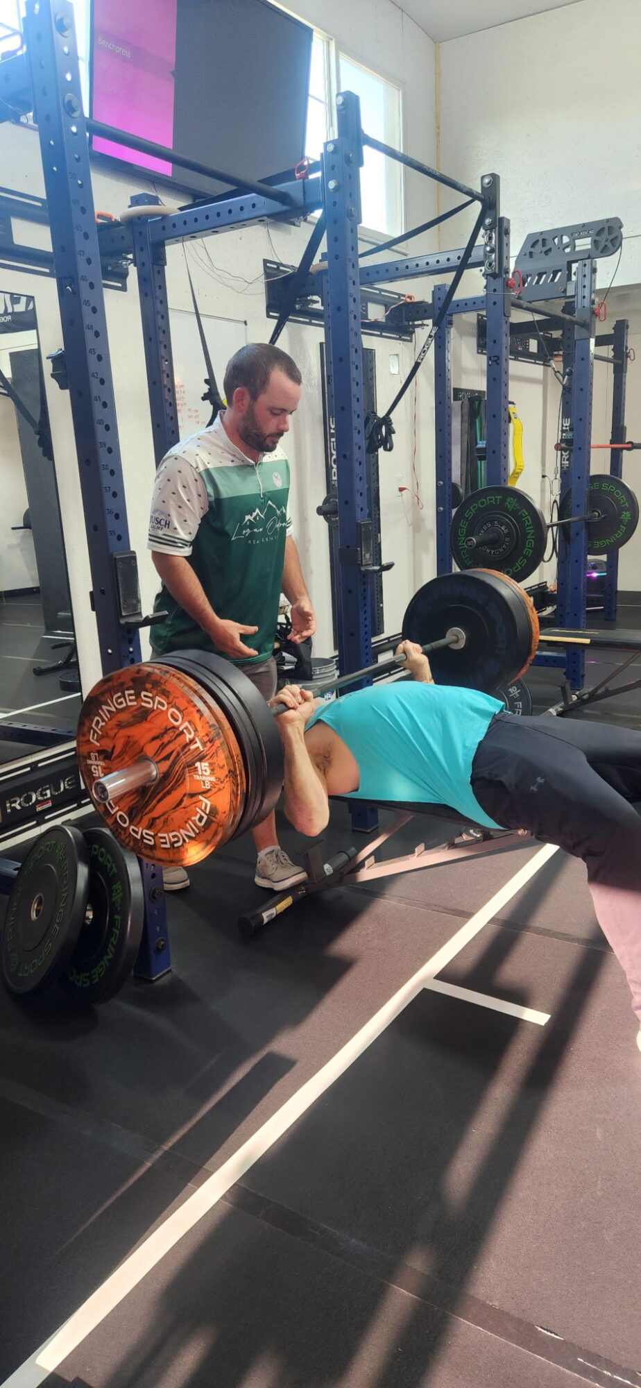 Person lying on a bench pressing a barbell with weights, spotter standing nearby in a gym.