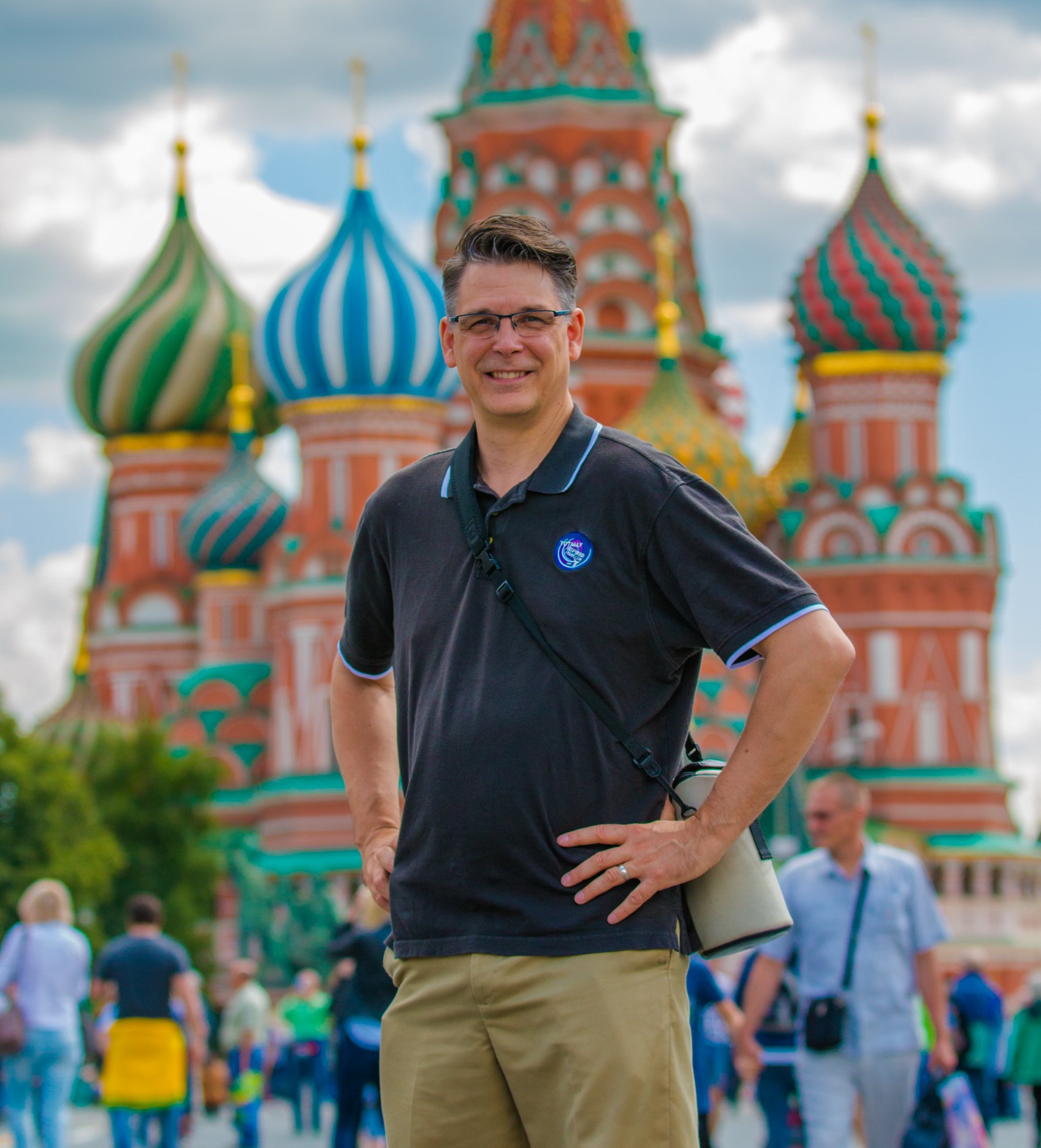 Smiling man in black polo shirt and khaki pants stands in front of colorful onion-domed building, with people in background.