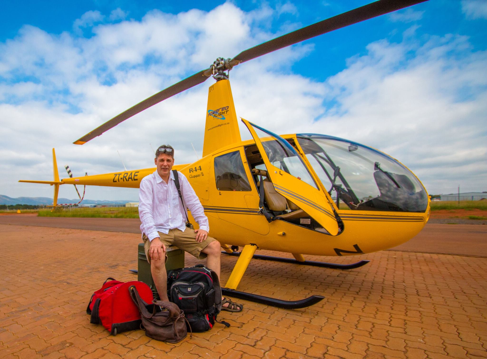 Person sitting on ground next to yellow helicopter with open door, backpacks nearby, on brick surface under blue sky.