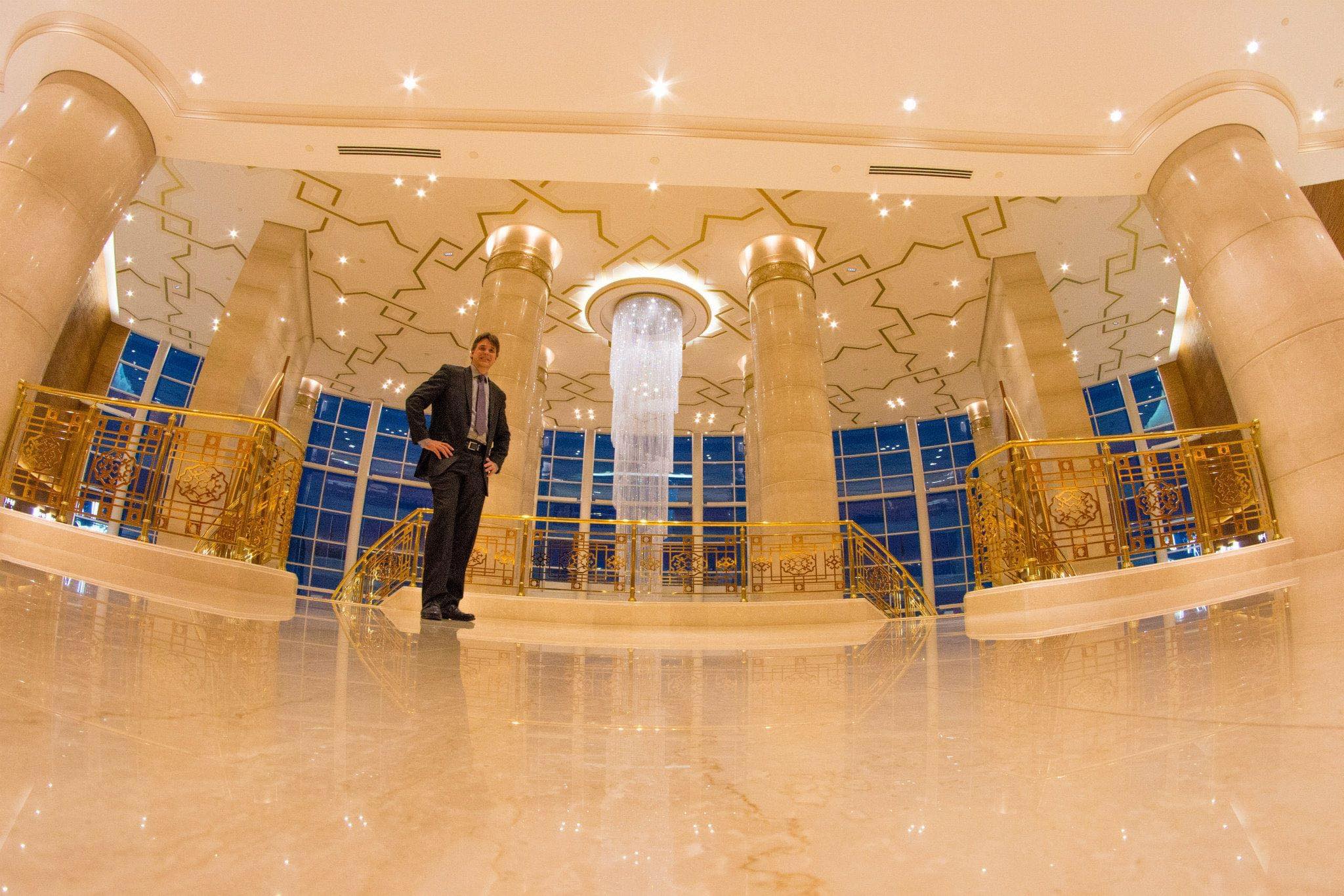 Man in suit standing in a grand, ornate hall with tall columns, gold accents, and a central illuminated feature.