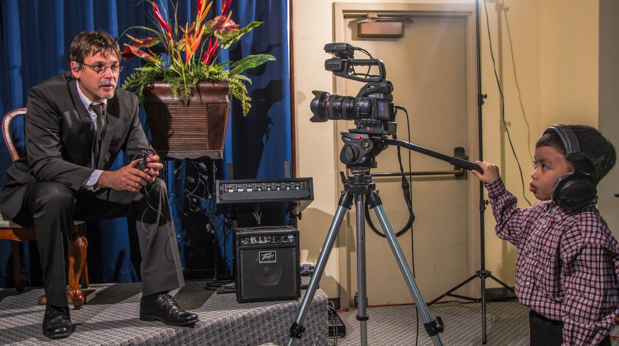 Child in headphones points a stick at a camera, man in suit sits nearby, recording equipment visible.