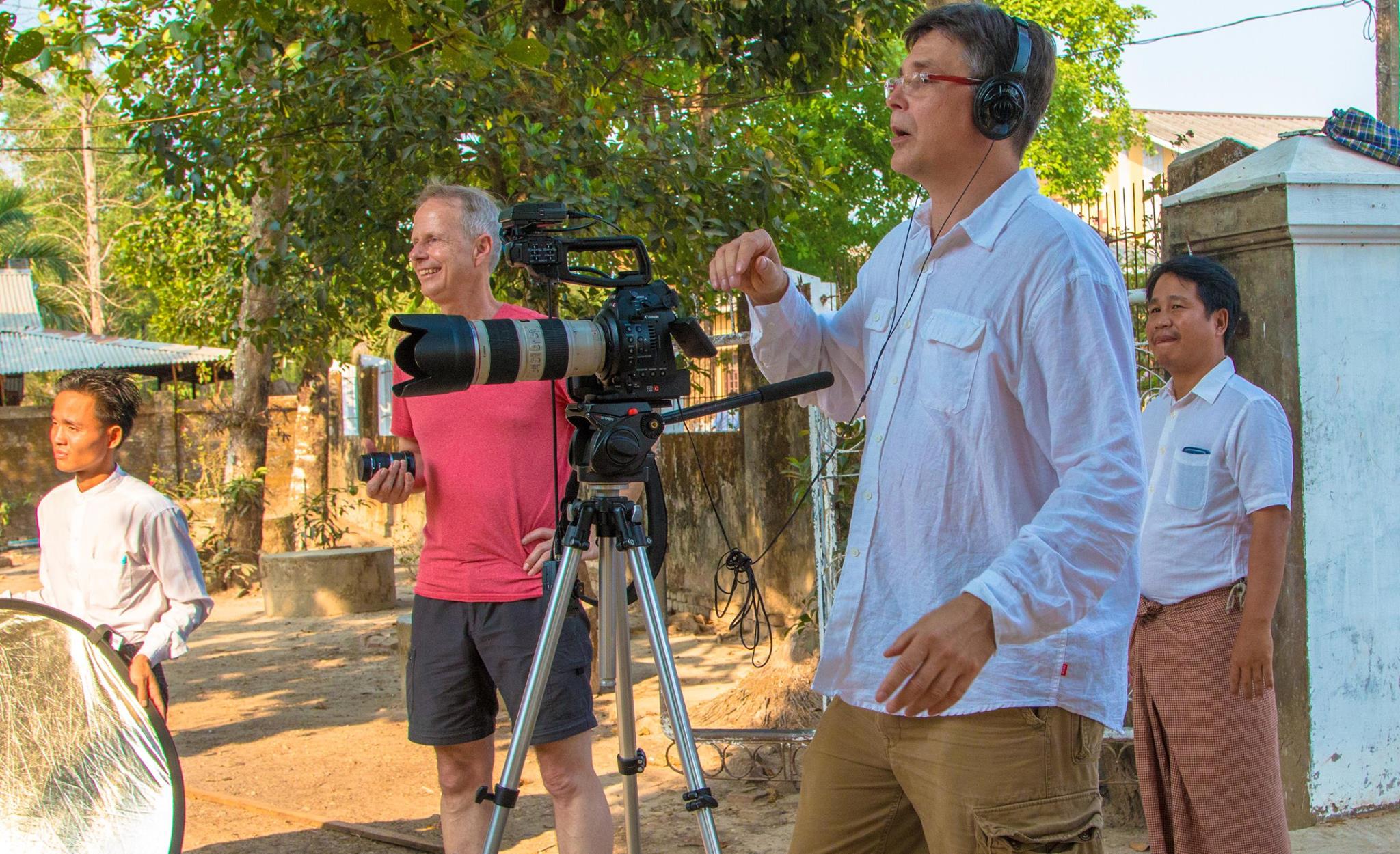 Group of people outdoors, one person with headphones operating a camera on a tripod, others smiling nearby.