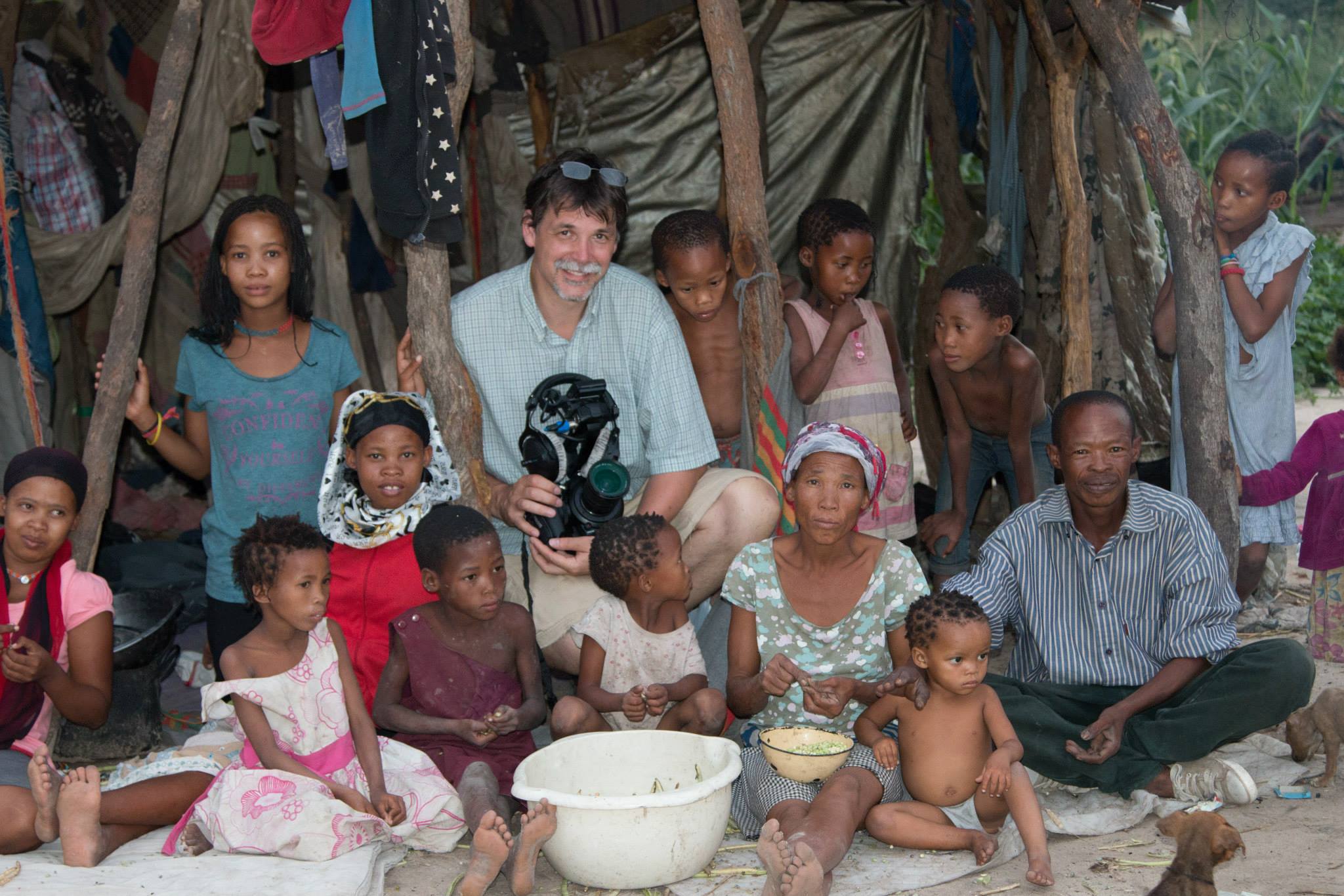 Group of children and adults gathered outdoors near a tent, some sitting and some standing, with a woman holding a camera.