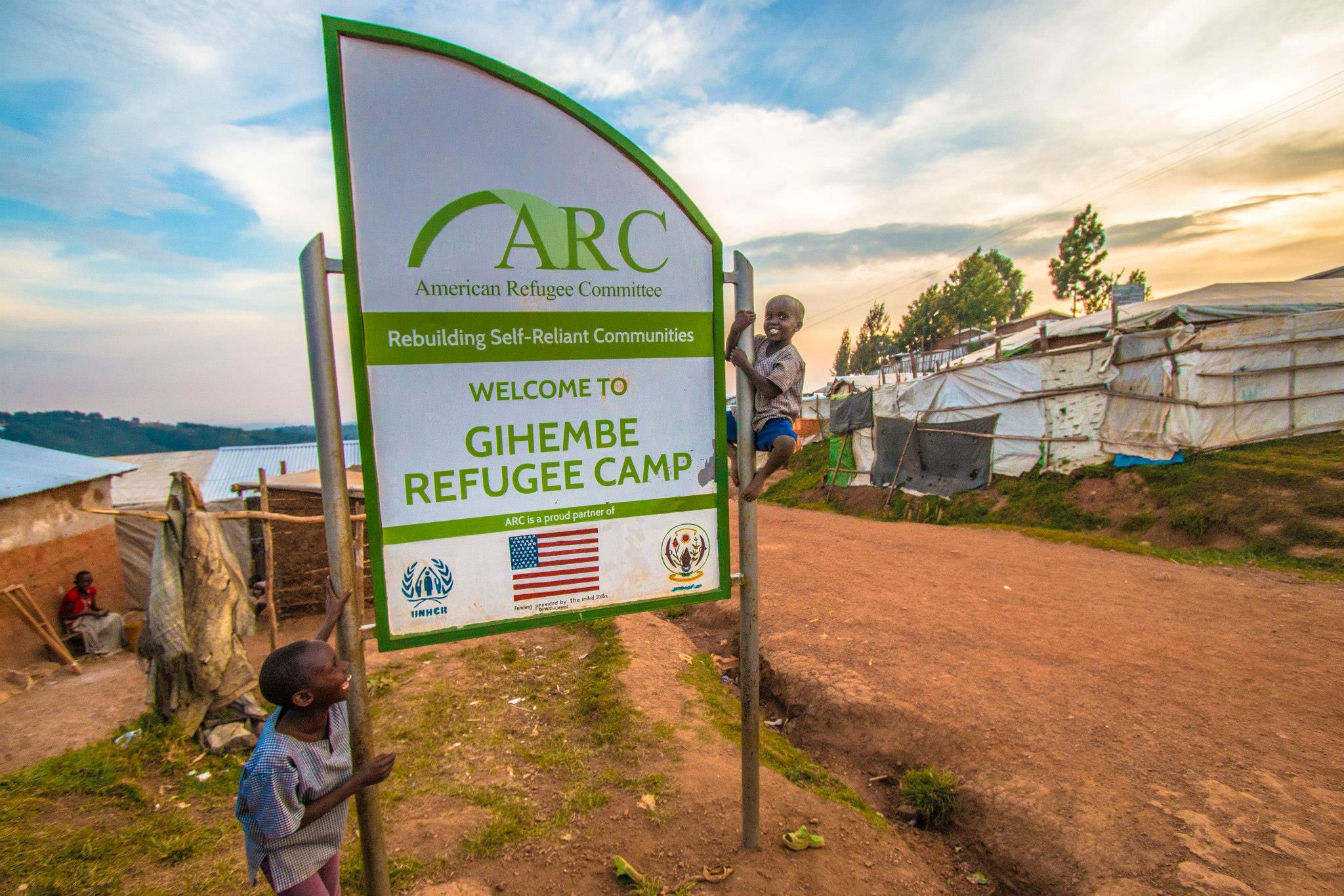 Signboard welcoming to Gihembe Refugee Camp with tents and trees in background.