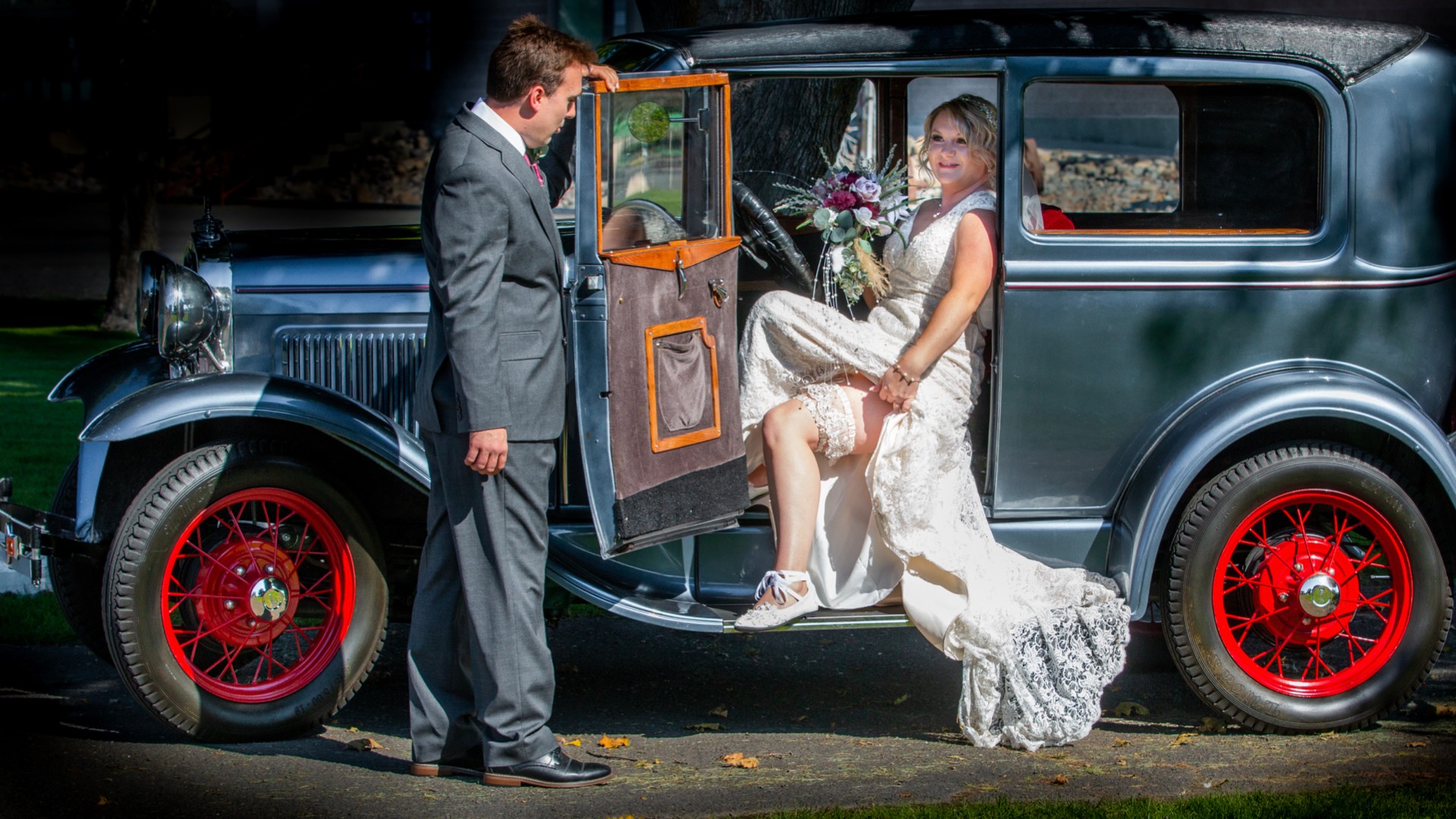 A bride in a wedding dress and veil sits in a vintage car, with a man in a suit standing outside.