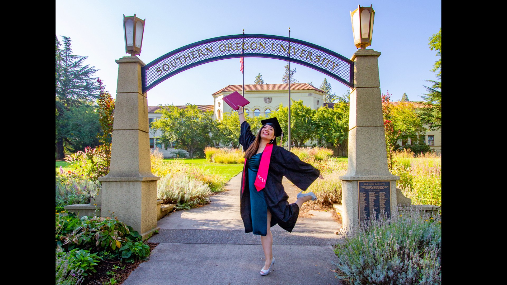 Graduate in cap and gown celebrating on campus pathway with university sign and buildings in background.