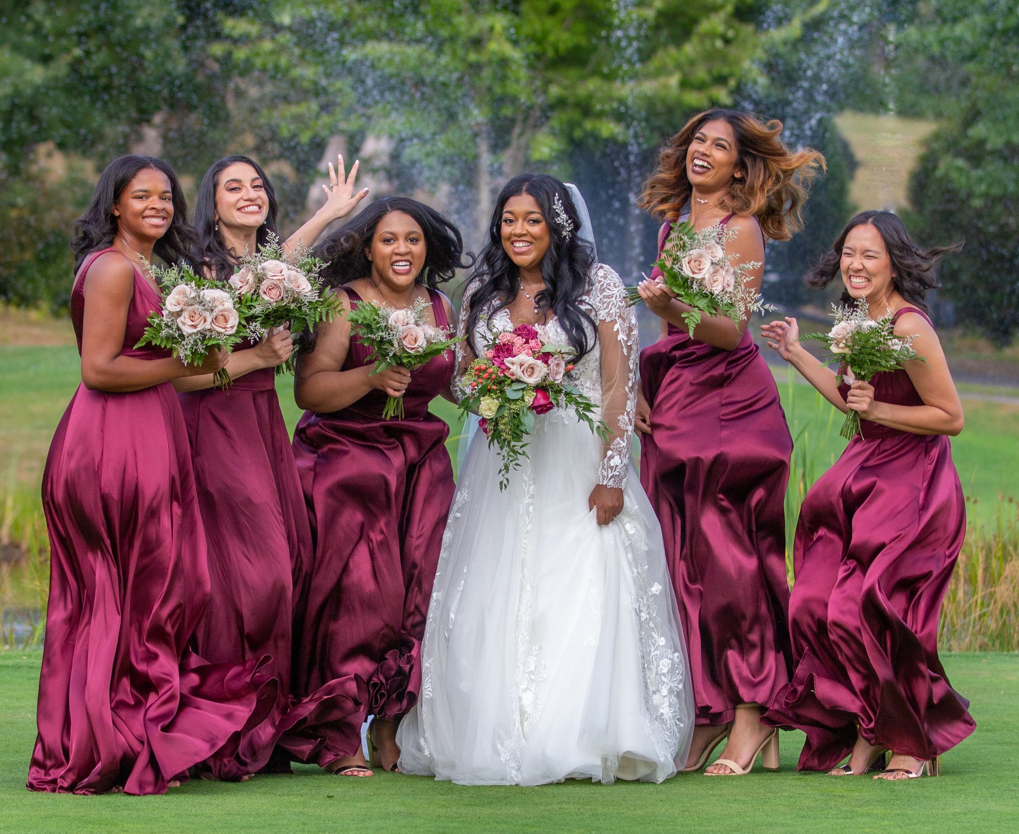 Group of six women, one in a white wedding dress, outdoors with trees, smiling and holding bouquets.