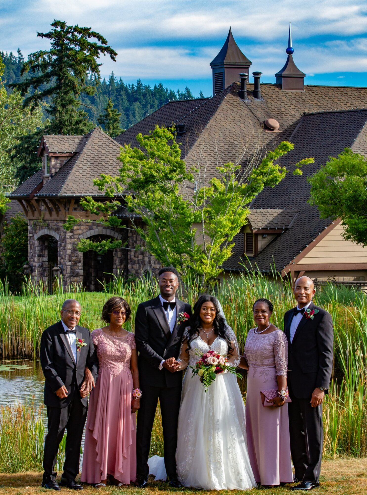 Group of six people, including a bride and groom, standing outdoors in front of a historic building with trees and a pond.