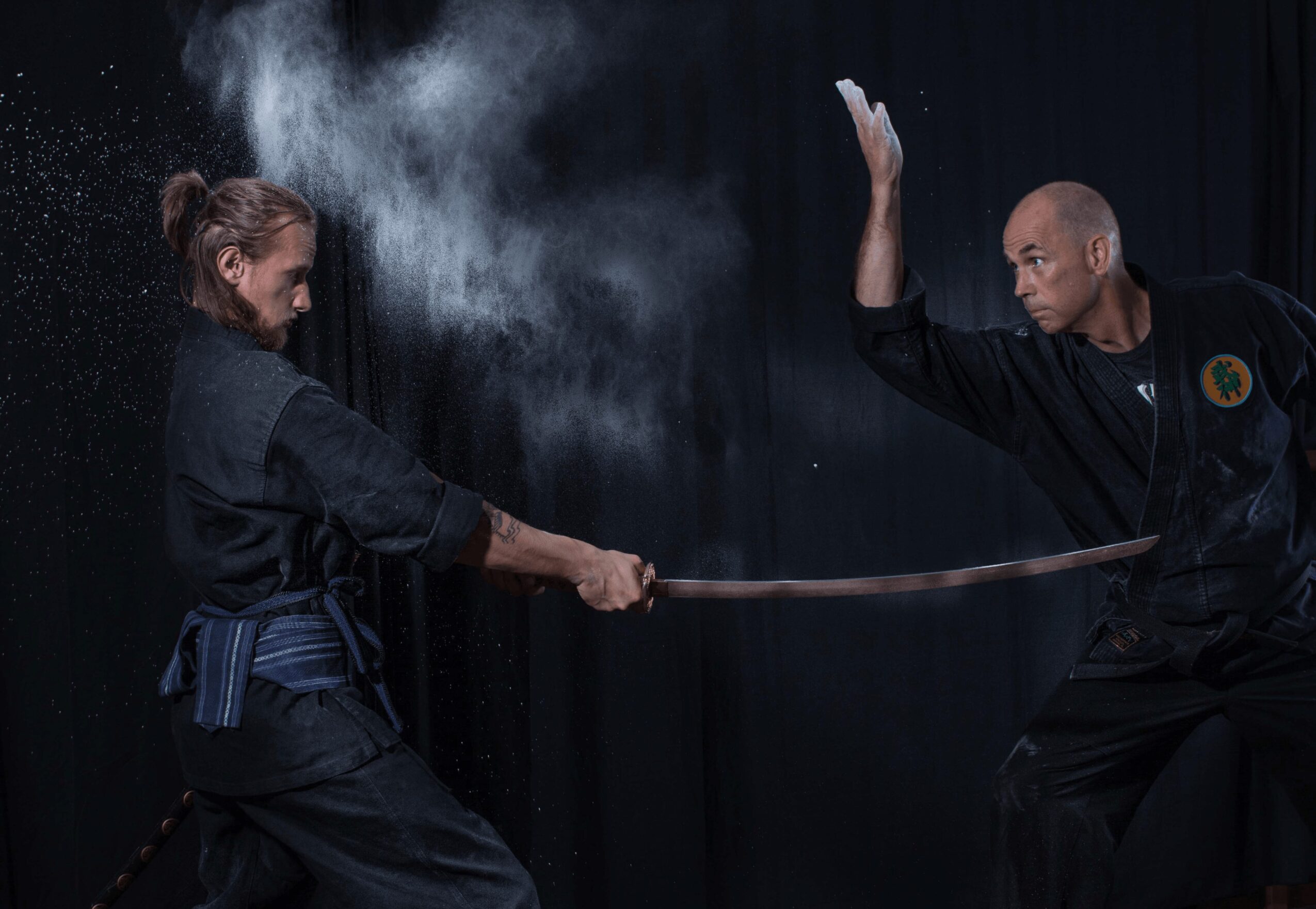 Two martial artists practicing with a sword, one releasing powder or dust, on a dark stage with a black curtain.