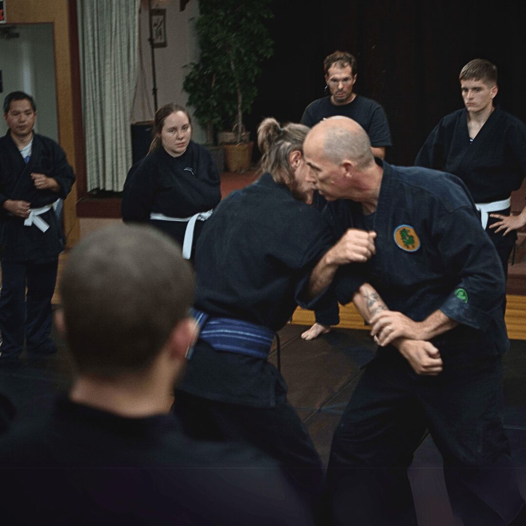 Two martial artists engage in a sparring match, surrounded by onlookers in a dojo setting.