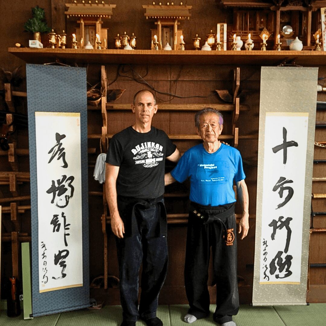 Two men standing side by side in front of Japanese calligraphy banners, inside a wooden room with shelves and decorations.