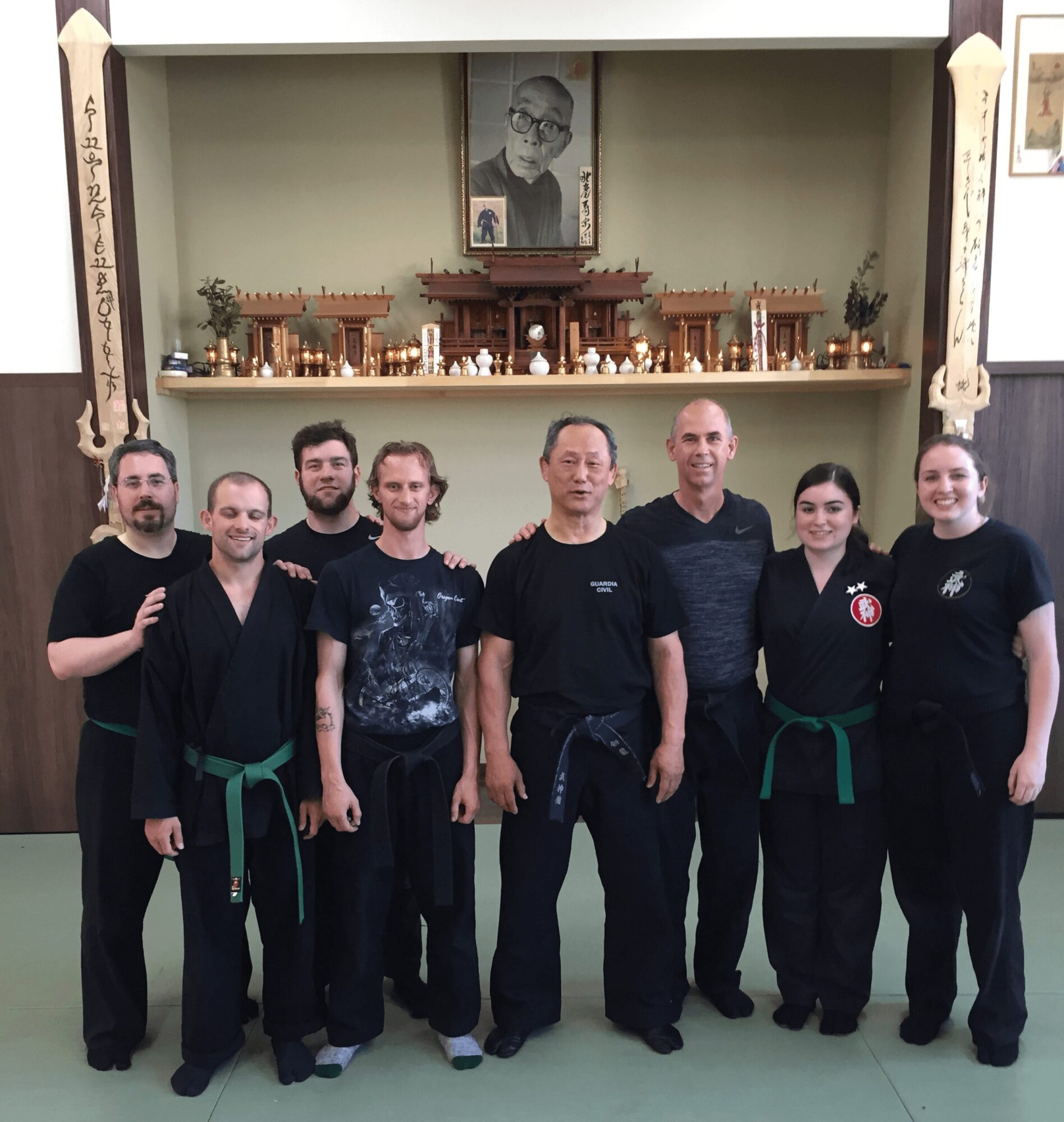 Group of seven people standing in front of an altar with a portrait and candles, some wearing martial arts uniforms with green belts.
