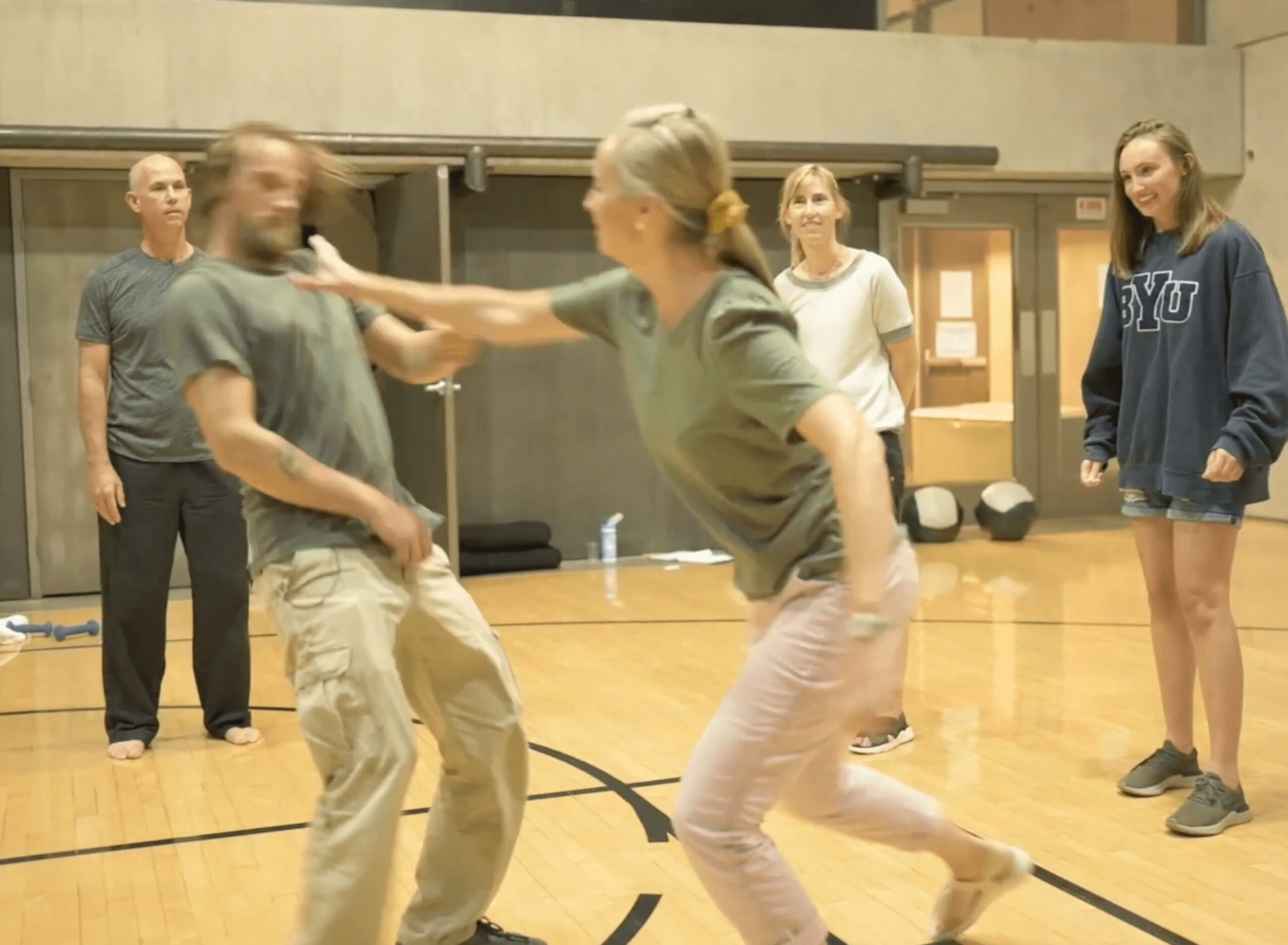 Group of people in a gym, with two women fighting and others watching, gym floor and walls visible.