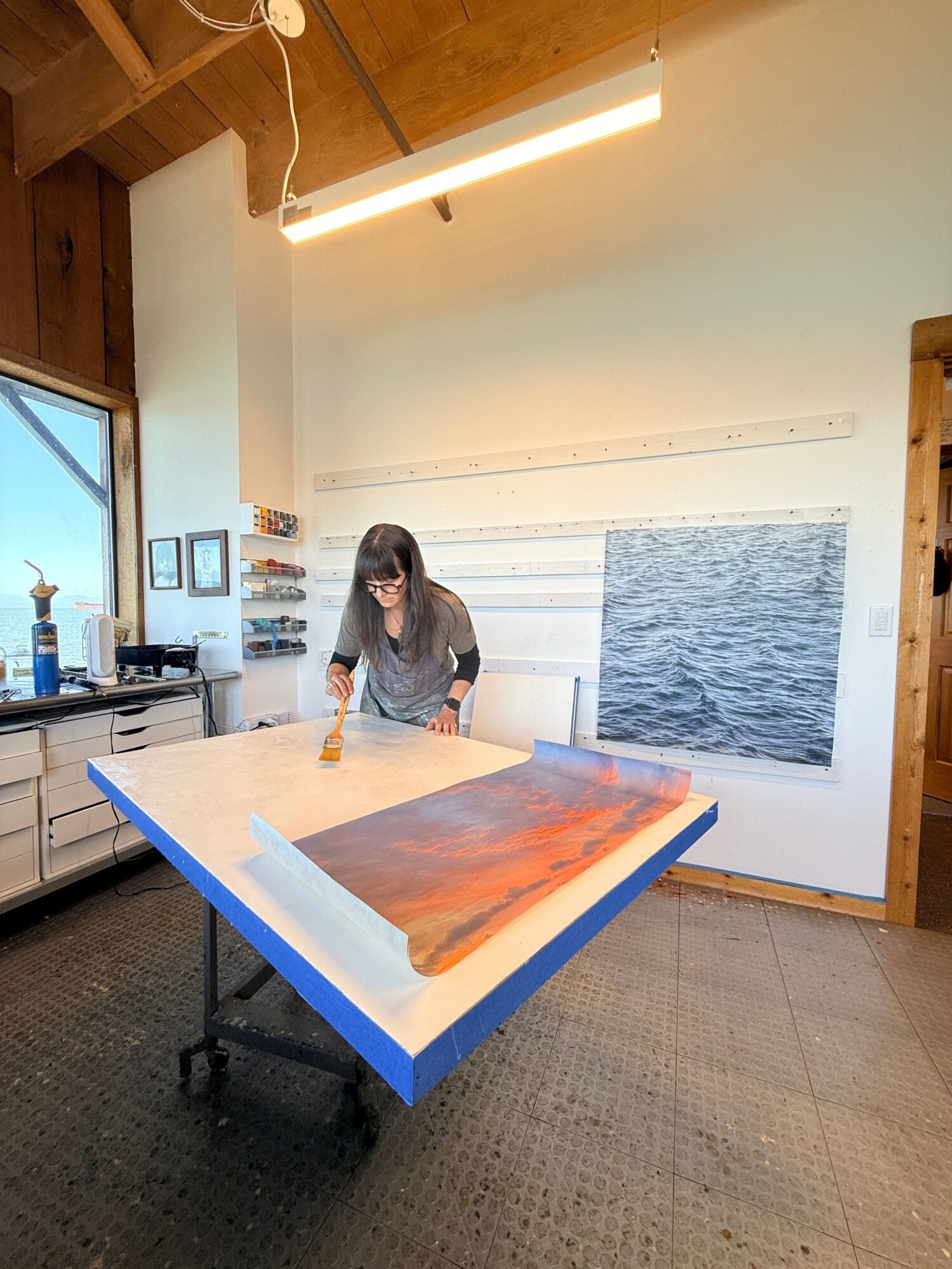 Person standing in a room with a large window, painting on a table, and shelves with jars in the background.