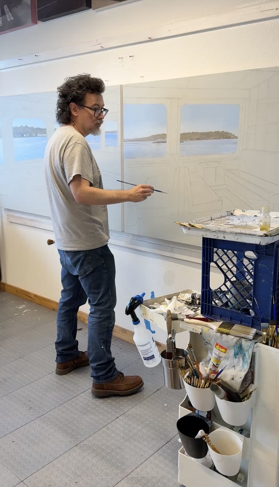 Man with curly hair and glasses painting landscape scenes on a wall, with art supplies nearby.