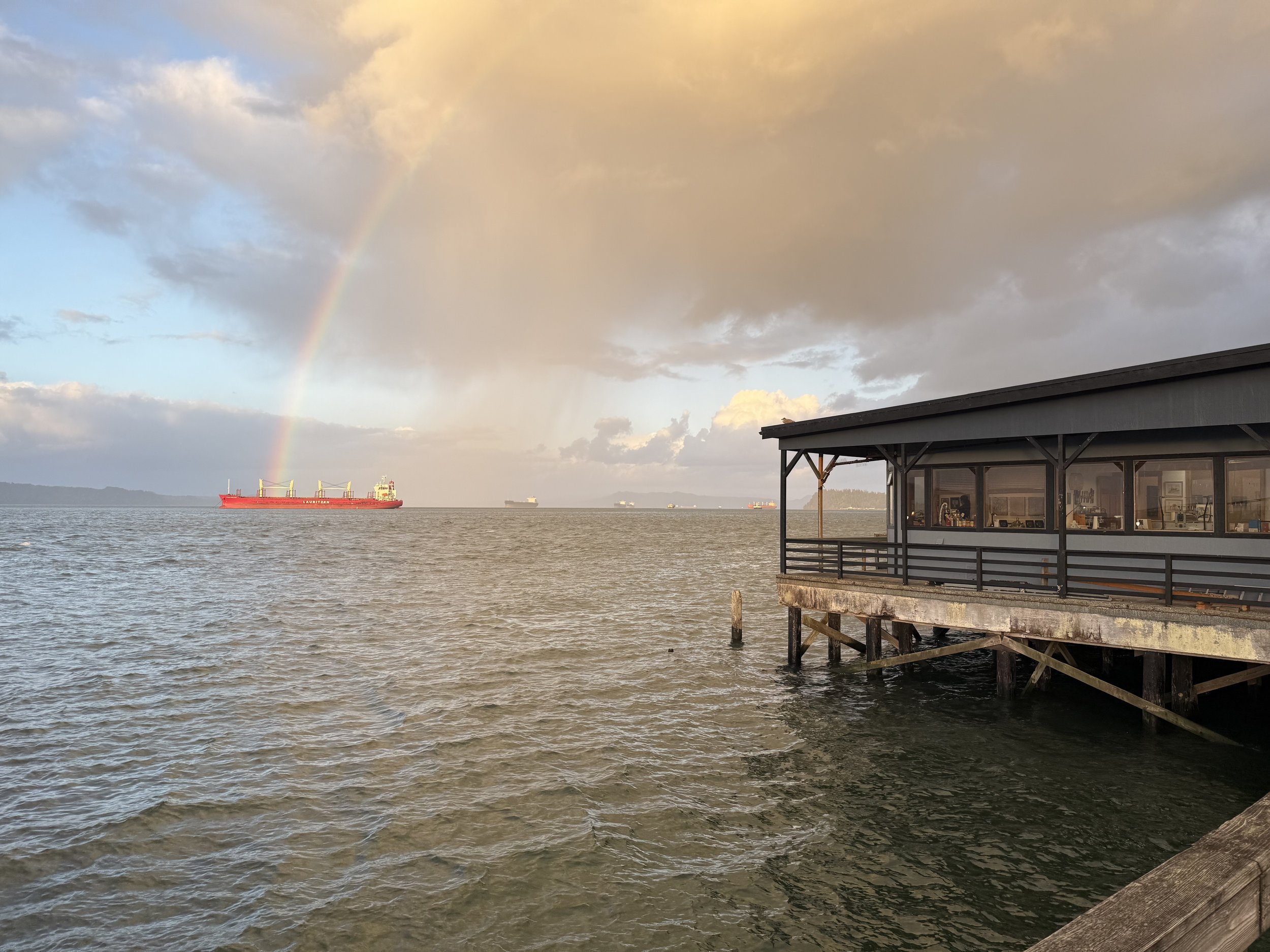 View of a body of water with a building on stilts, a ship in the distance, and a rainbow in the sky.