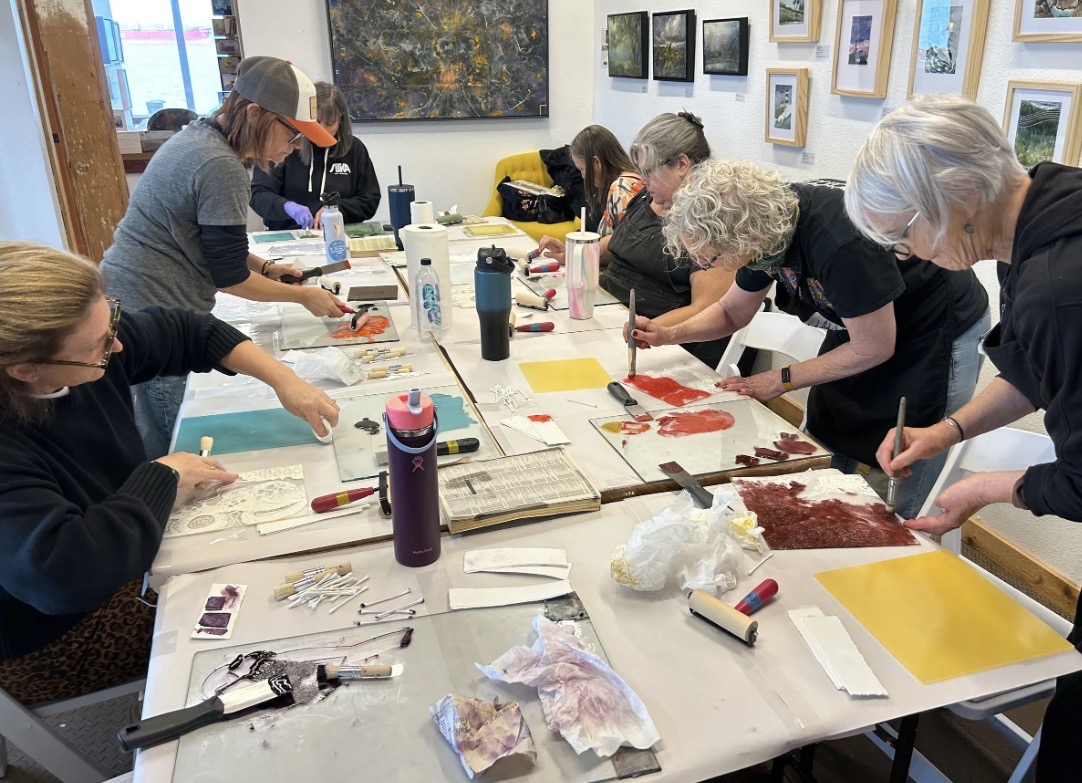 Group of people working on art projects at a table in a room with framed pictures on the wall.