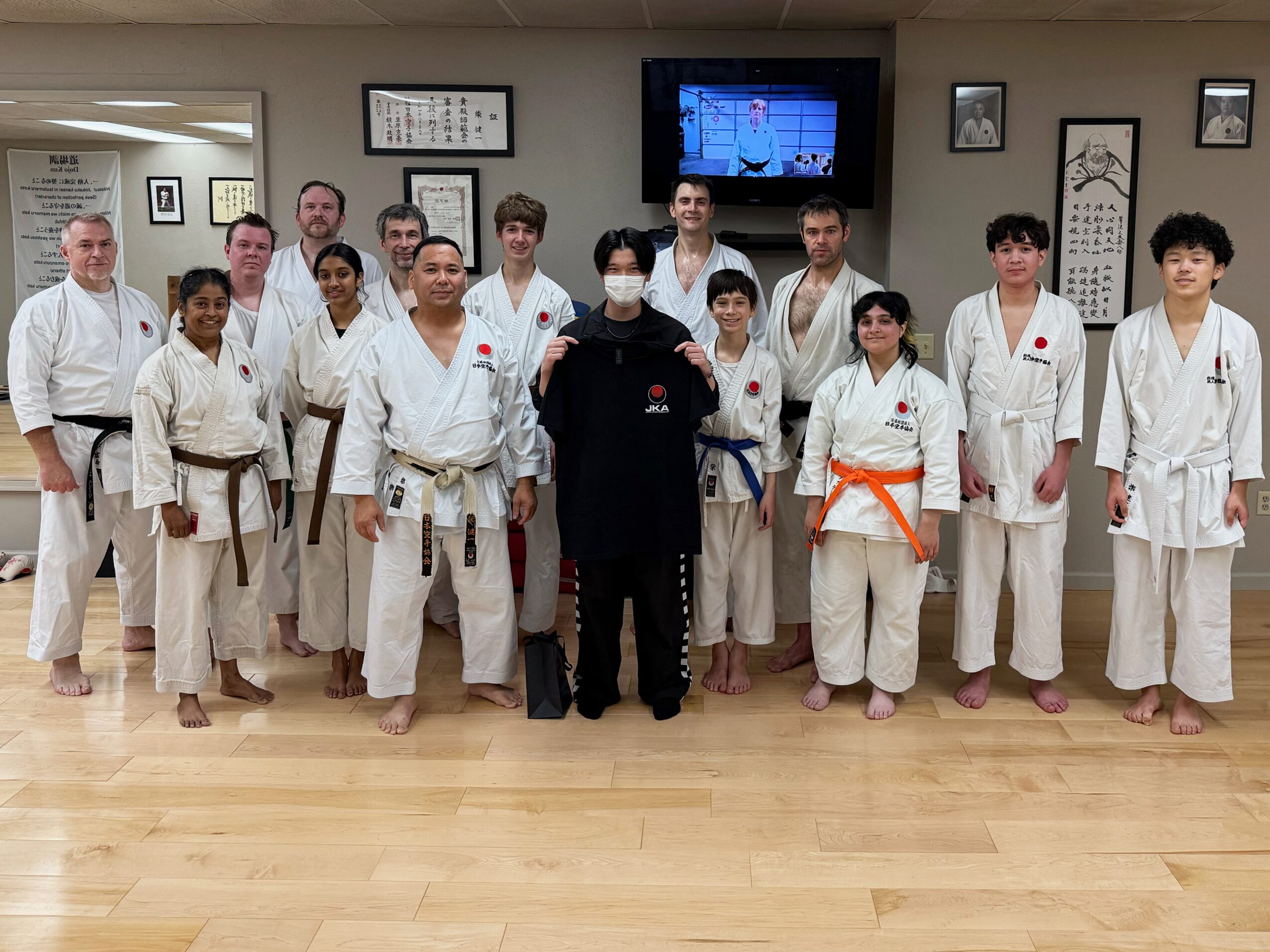 Group of people in martial arts uniforms standing in a dojo, some with belts of different colors, with a person in black in the center.