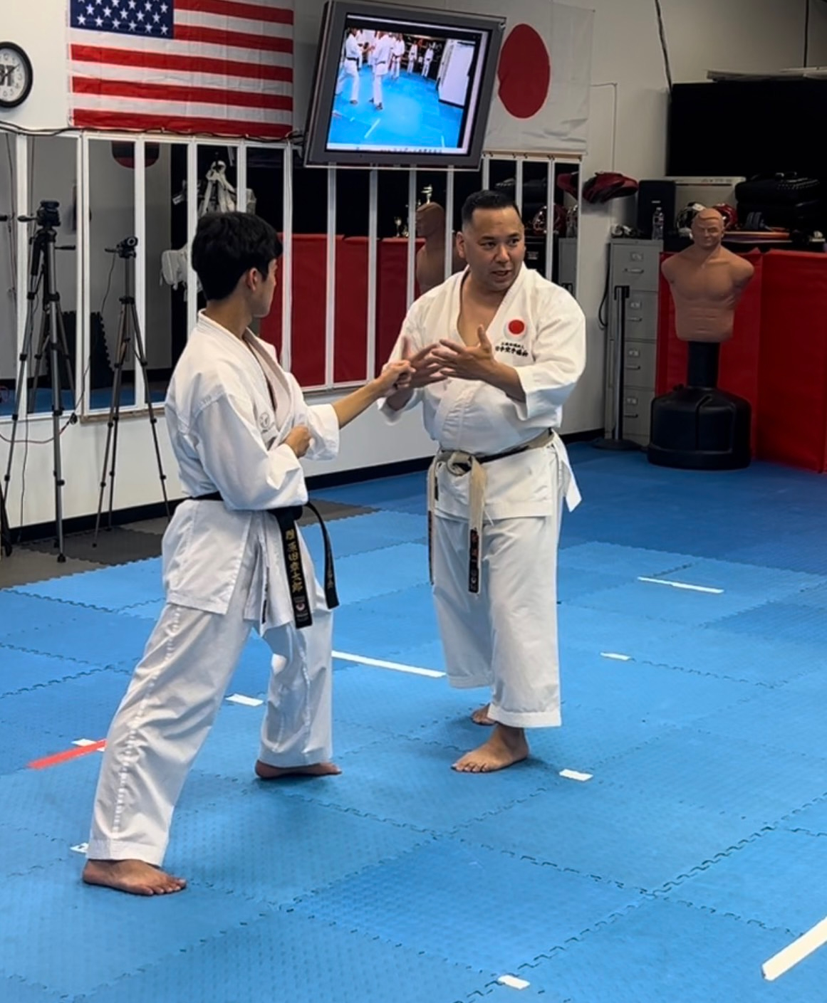 Two martial artists practicing in a dojo, one demonstrating a technique to the other, with flags and training equipment in background.
