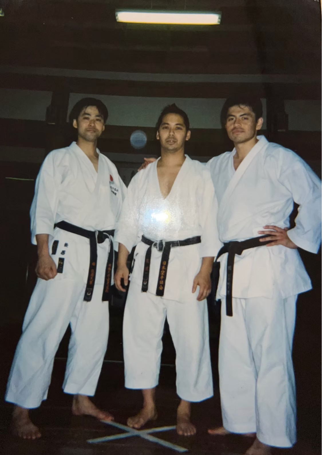 Three people in martial arts uniforms standing together indoors, with a dark background and a light fixture above. All wear black belts.