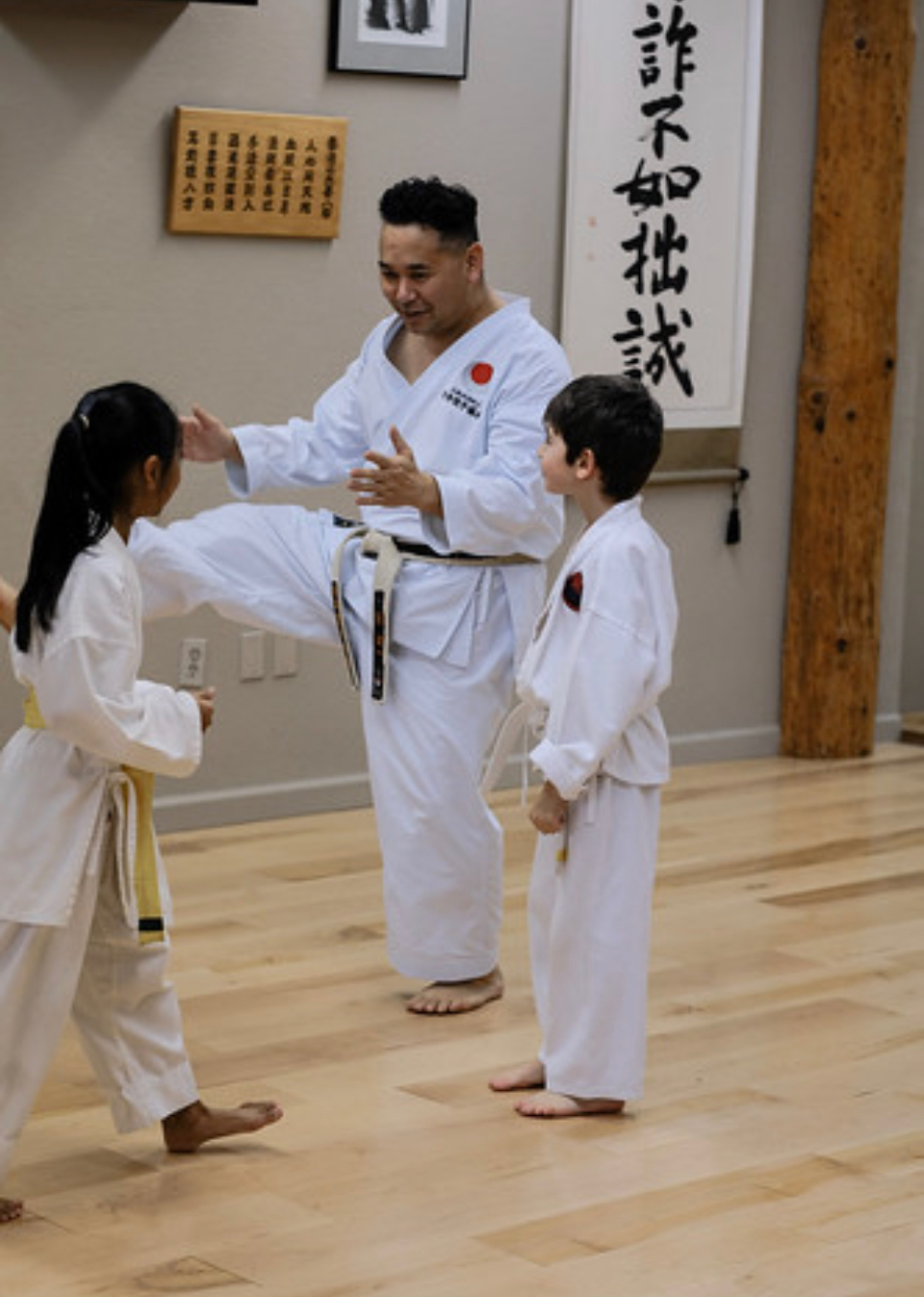 Martial arts instructor demonstrating techniques to two children in a dojo, with Japanese calligraphy on the wall.