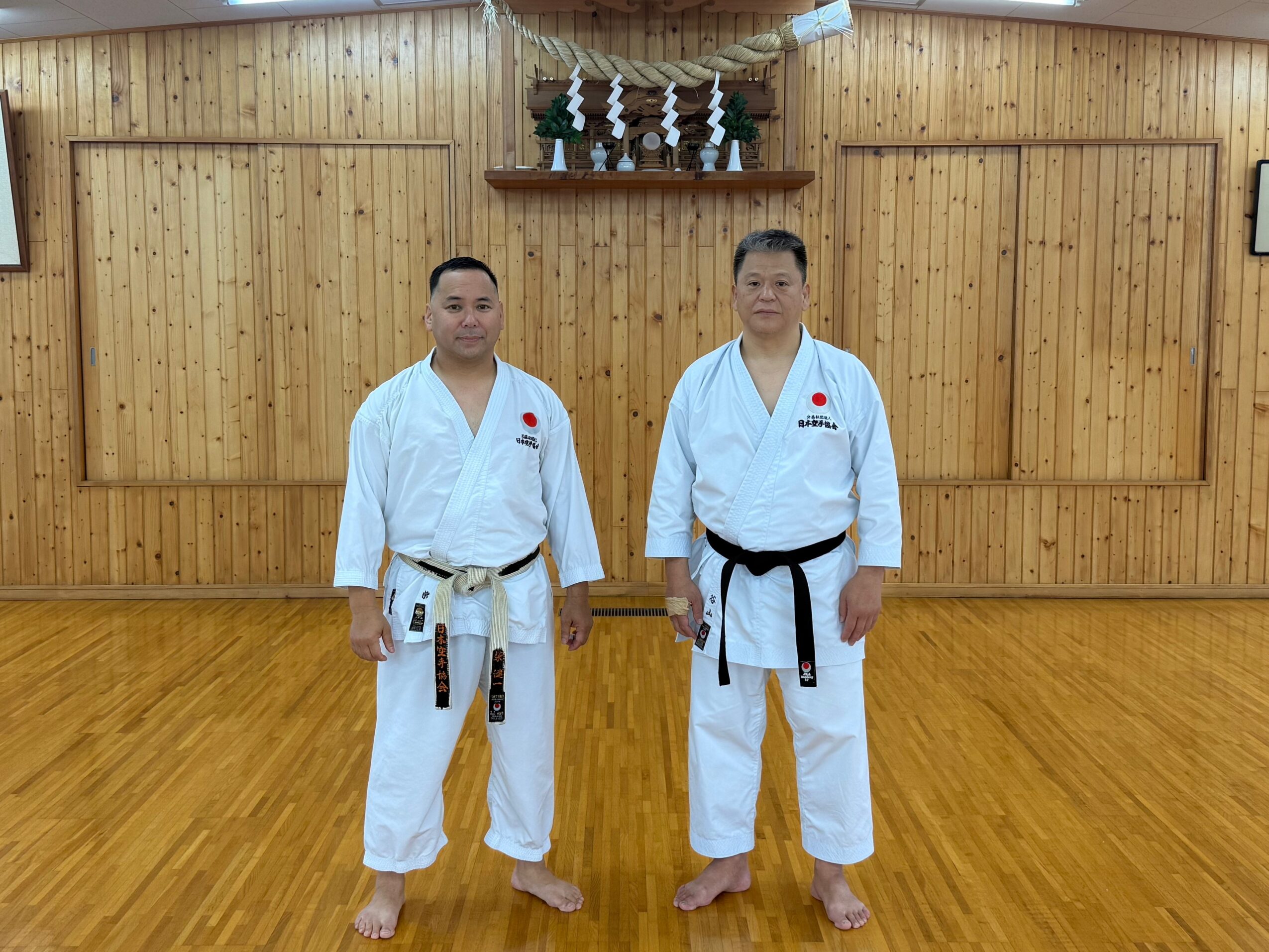 Two people in martial arts uniforms stand in a wooden dojo, facing forward with hands at sides, wooden wall background.