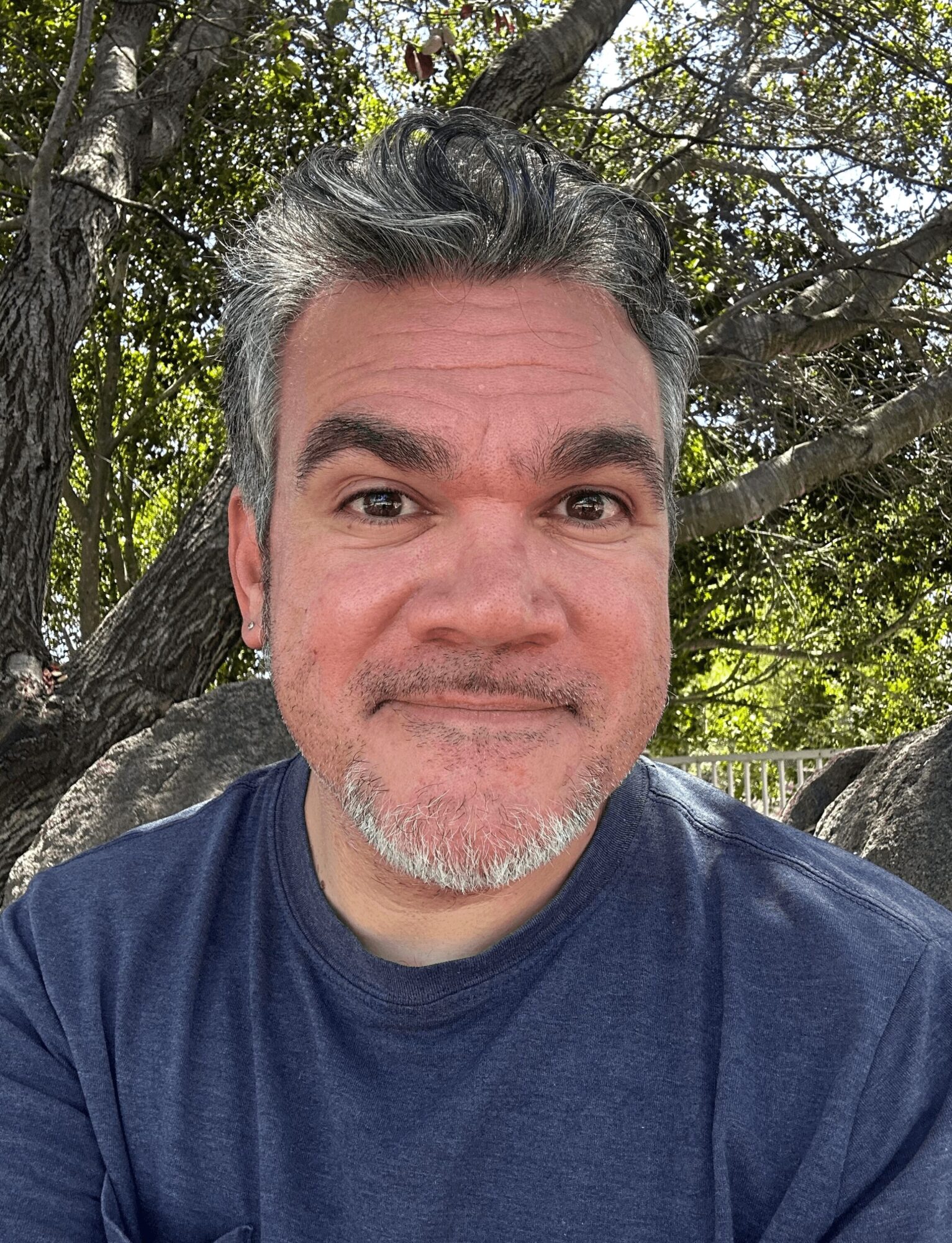 Man with gray hair and beard smiling outdoors with trees in the background.