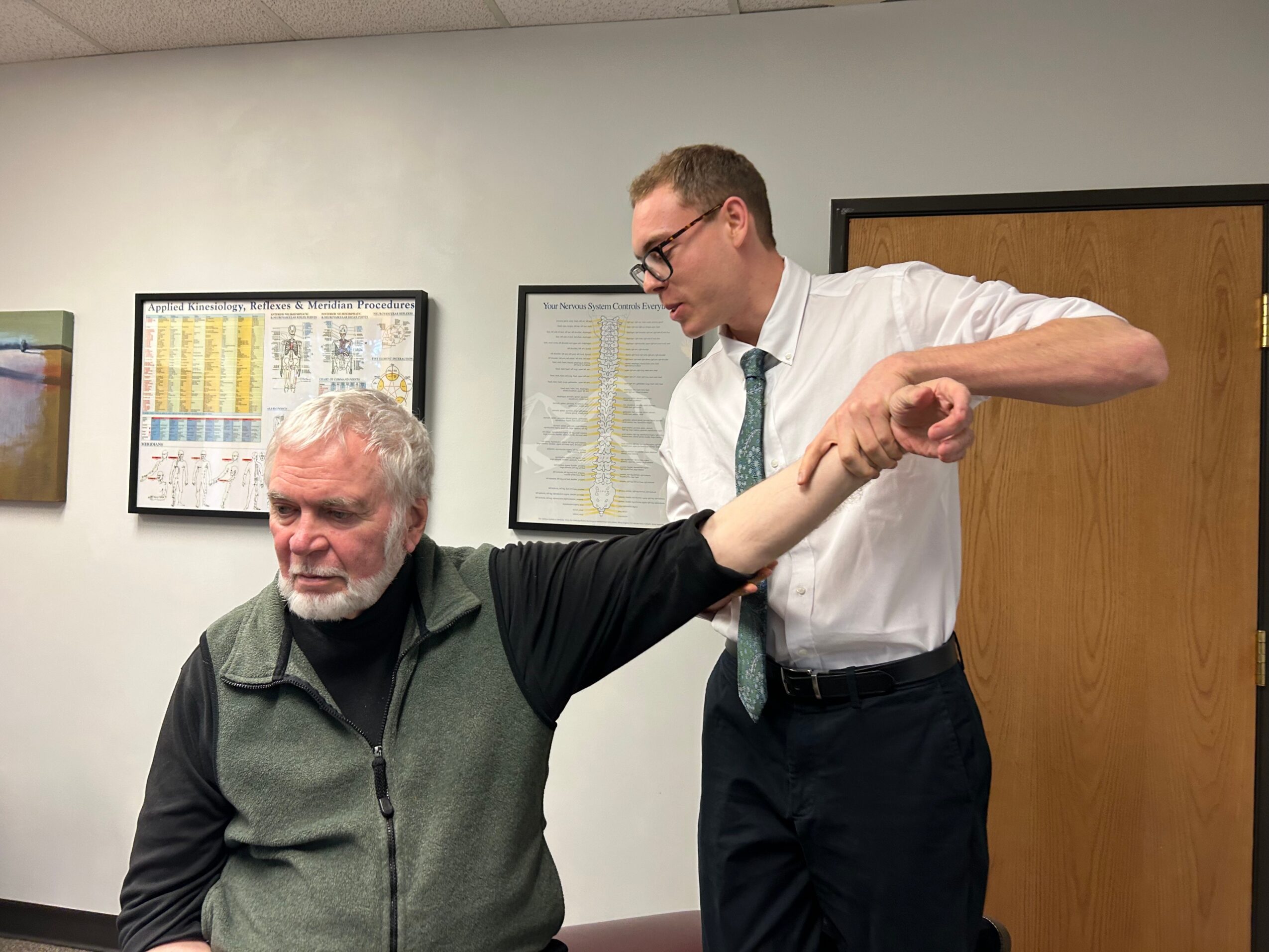 A man with glasses and a white shirt is assisting an older man with a beard, who is wearing a black and green vest, in an office setting.