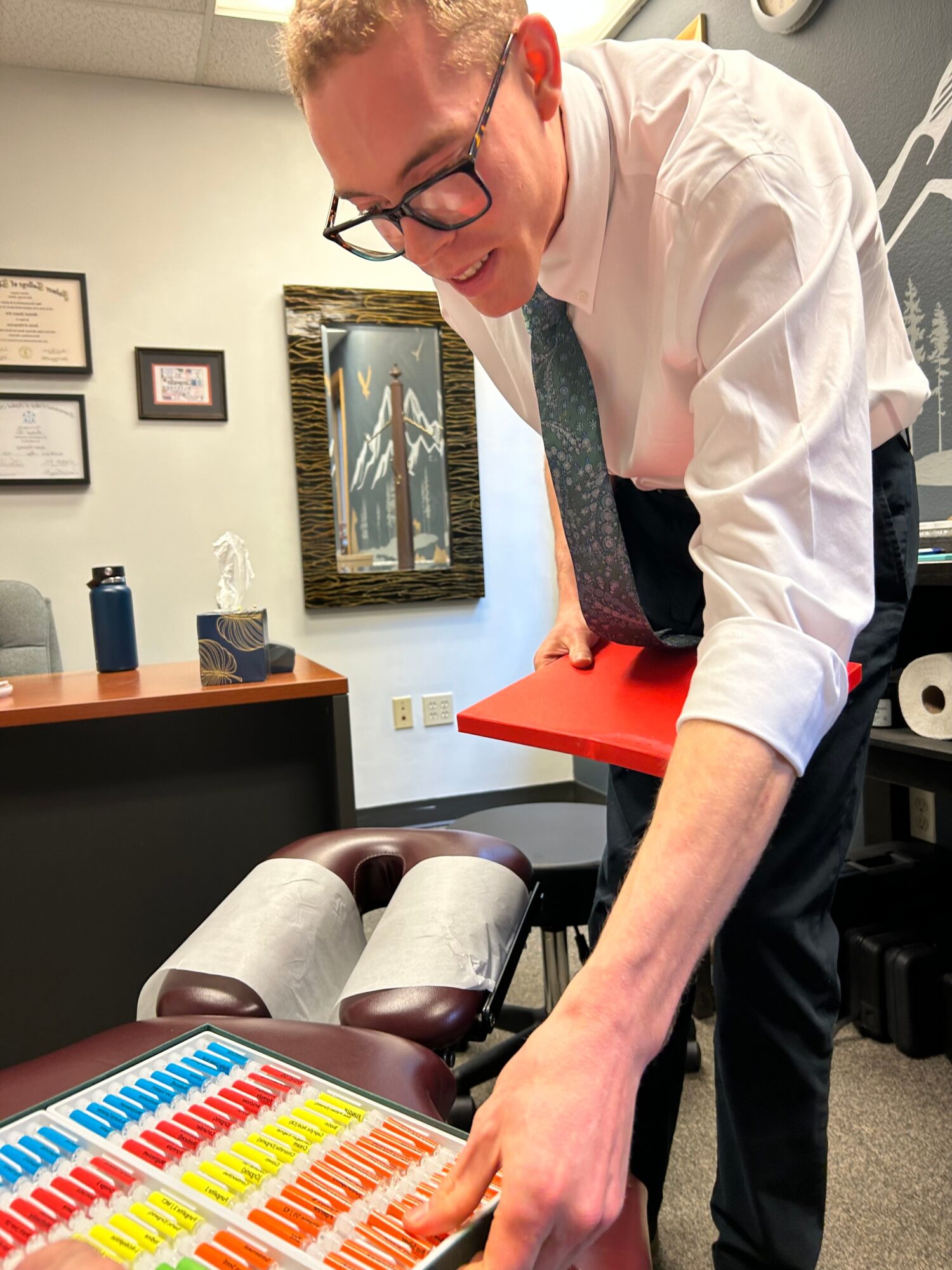Man in white shirt and tie leaning over a table with colorful objects, in an office setting.