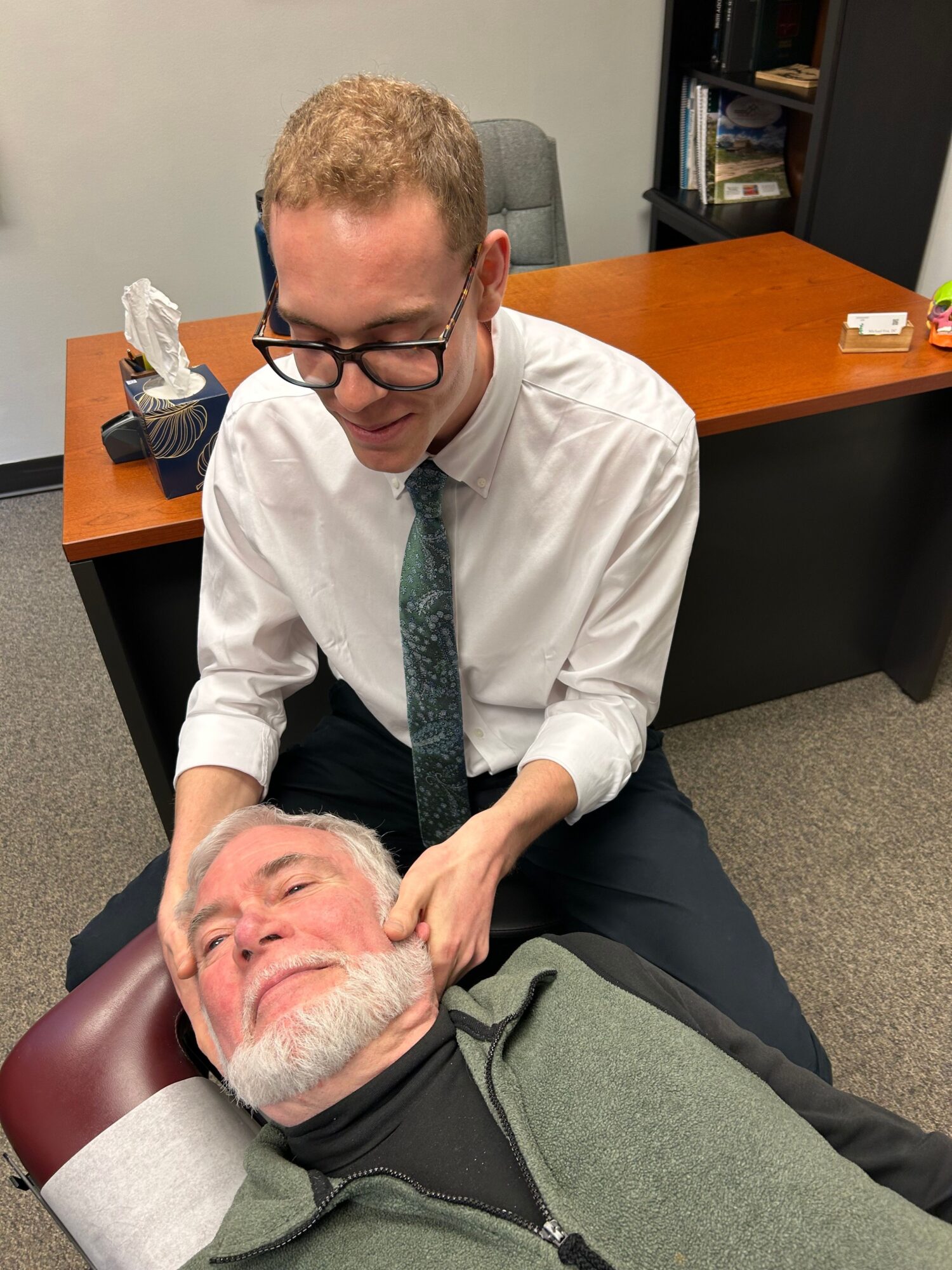 Two men in an office, one lying on a chair, the other leaning over, appearing to check the other's pulse or health.