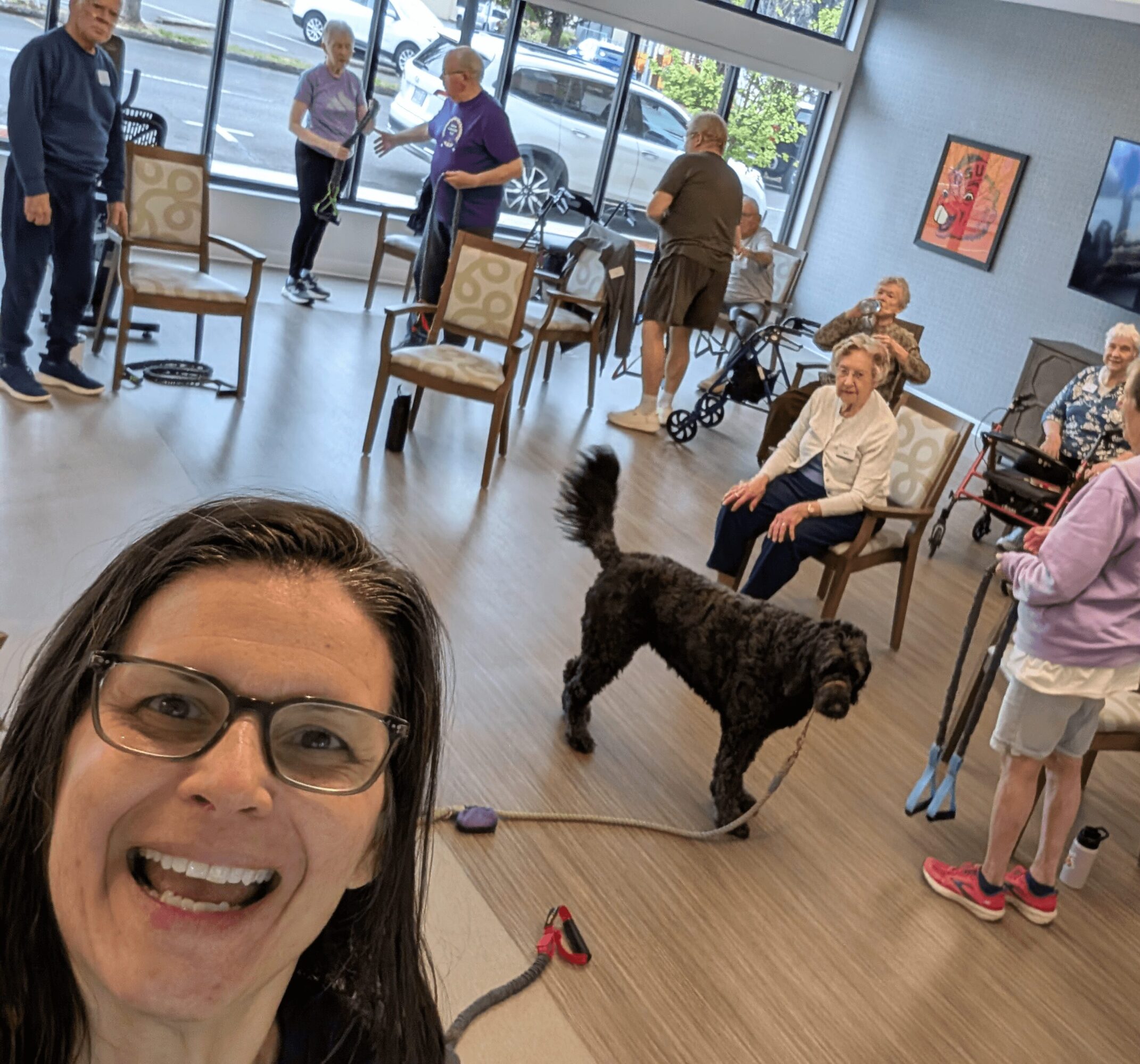 Woman with glasses smiling in foreground, people and a dog in a room with large windows and chairs.
