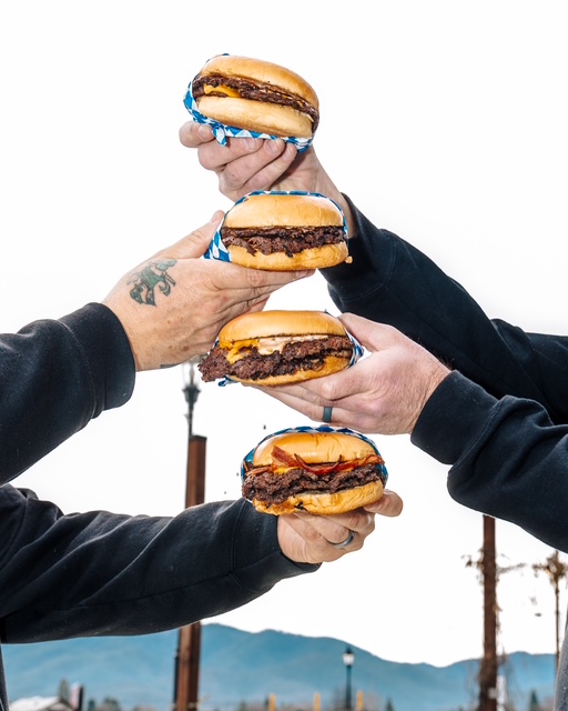 Four hands holding hamburgers stacked vertically against a background of mountains and sky.
