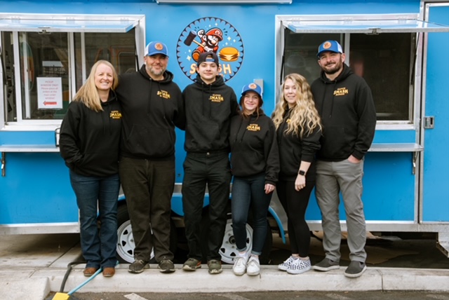 Group of six people standing in front of a blue food truck, smiling, wearing black hoodies and caps.
