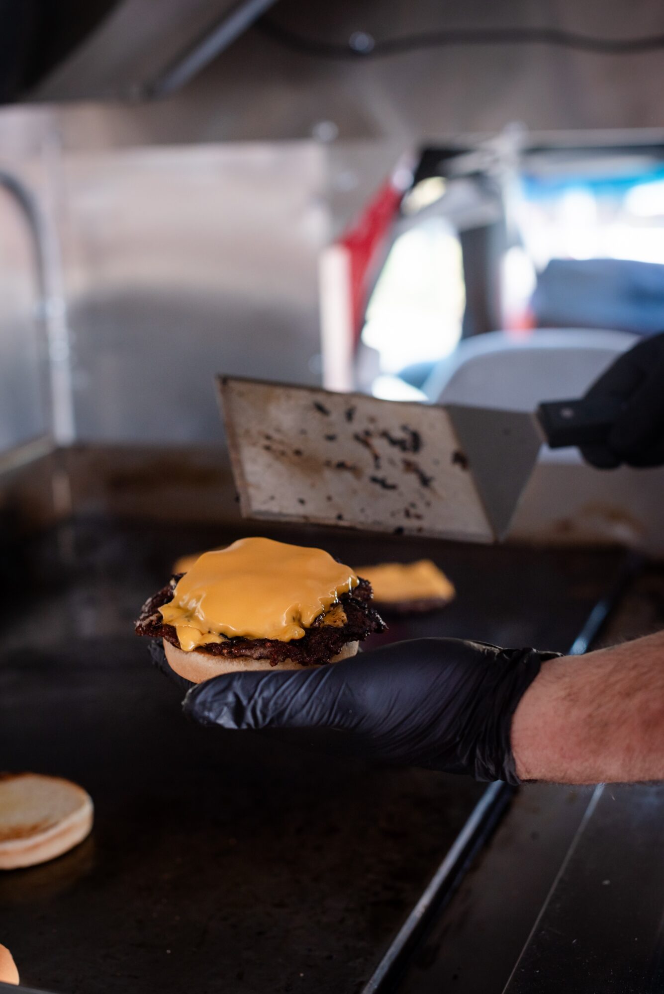 Person wearing black glove preparing a cheeseburger on a flat-top grill, with a spatula in the background.