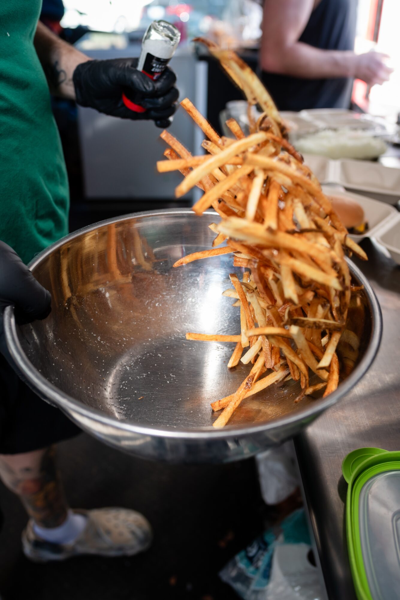Person pouring cooked French fries into a large metal bowl at a food stand, with other people in the background.