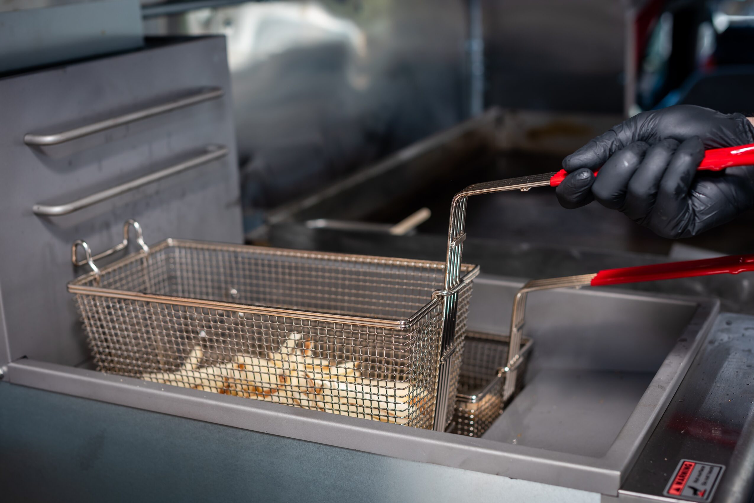 Person wearing black gloves uses tongs to handle food in a fryer basket, with a commercial fryer in the background.