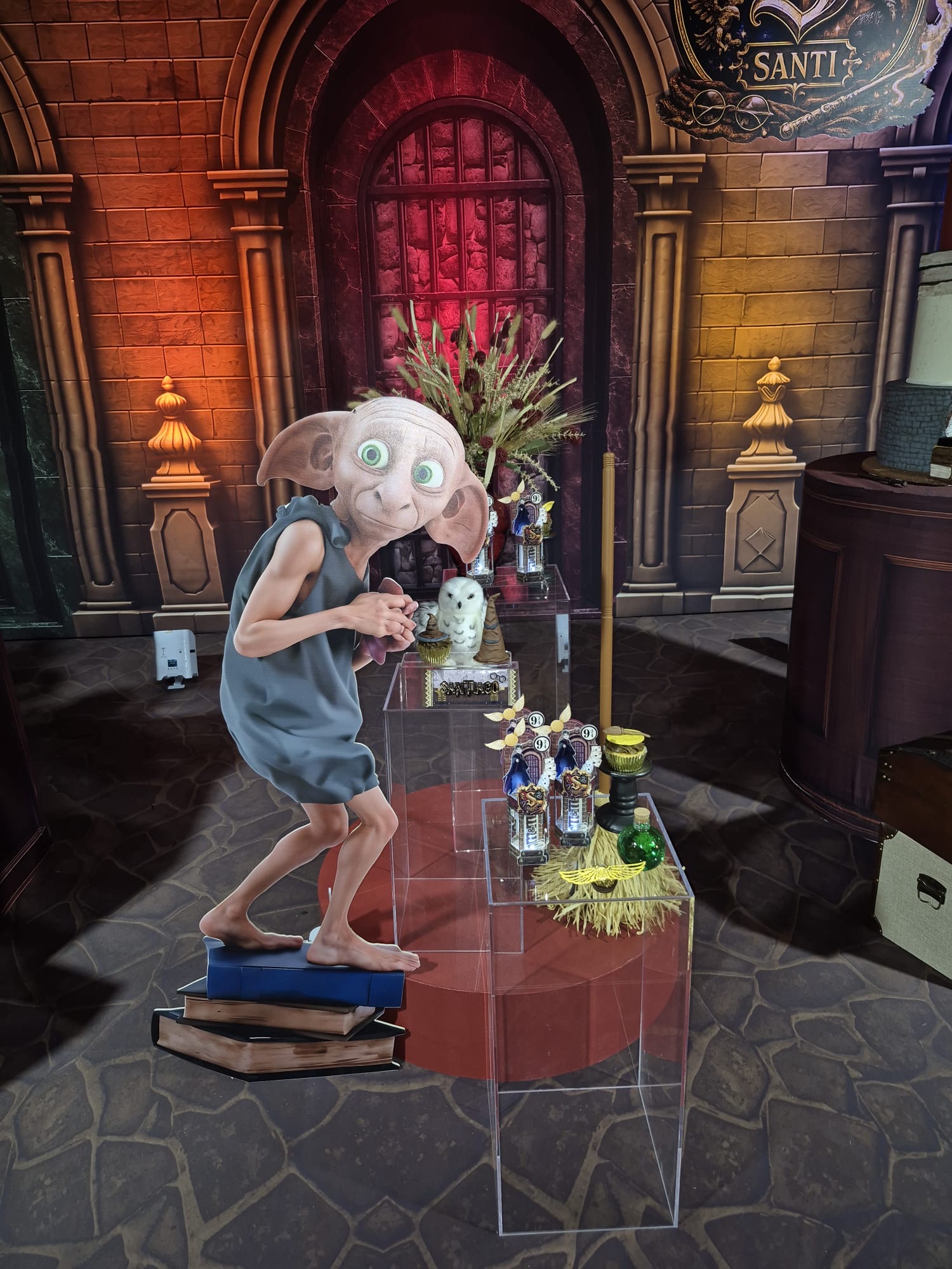 Person in a costume with large eyes and gray hair stands on a platform in a decorated room with plants and props.
