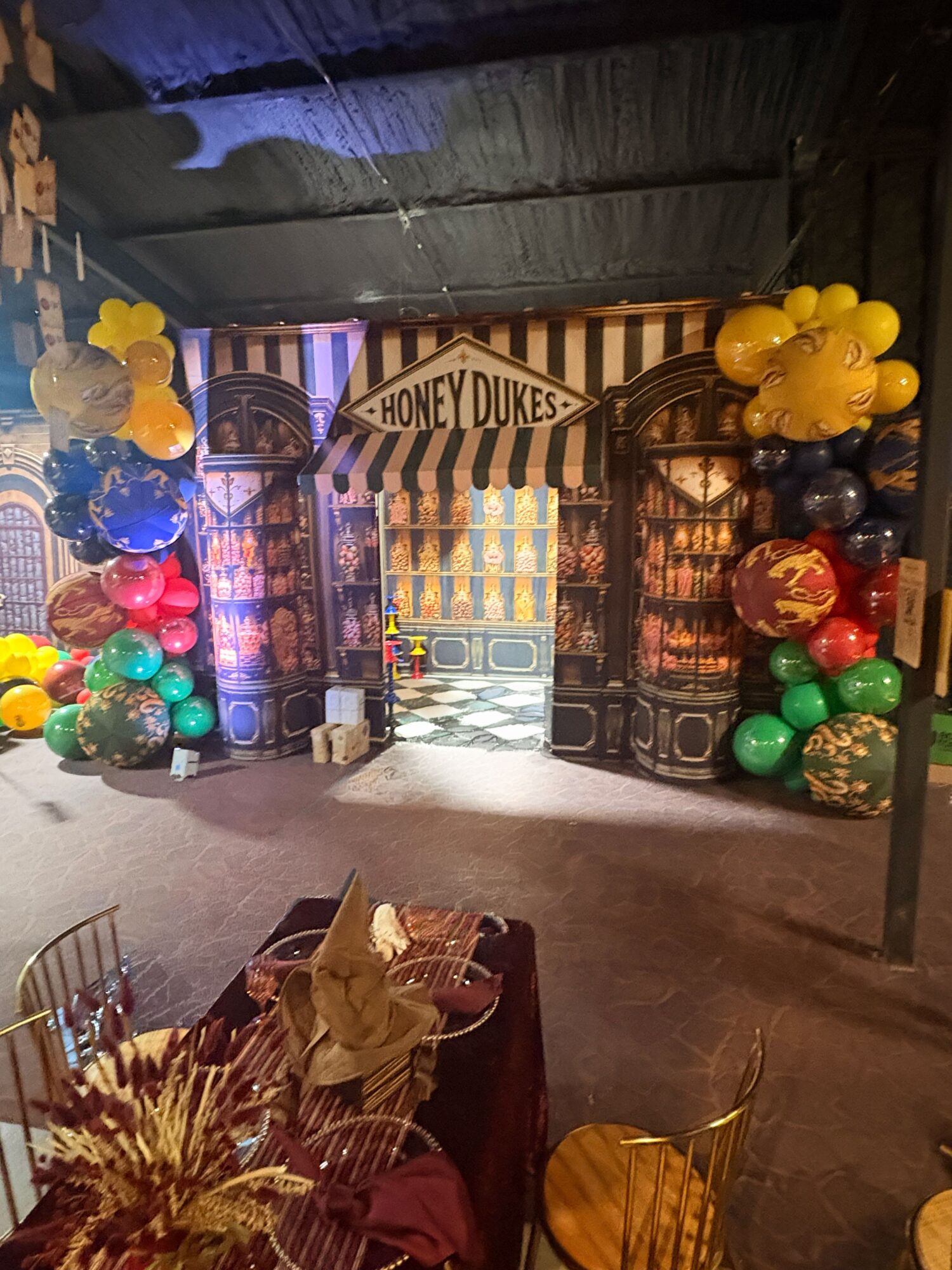 Decorated entrance with balloons and a sign reading 'Honey Dukes,' resembling a whimsical shop, with tables in foreground.